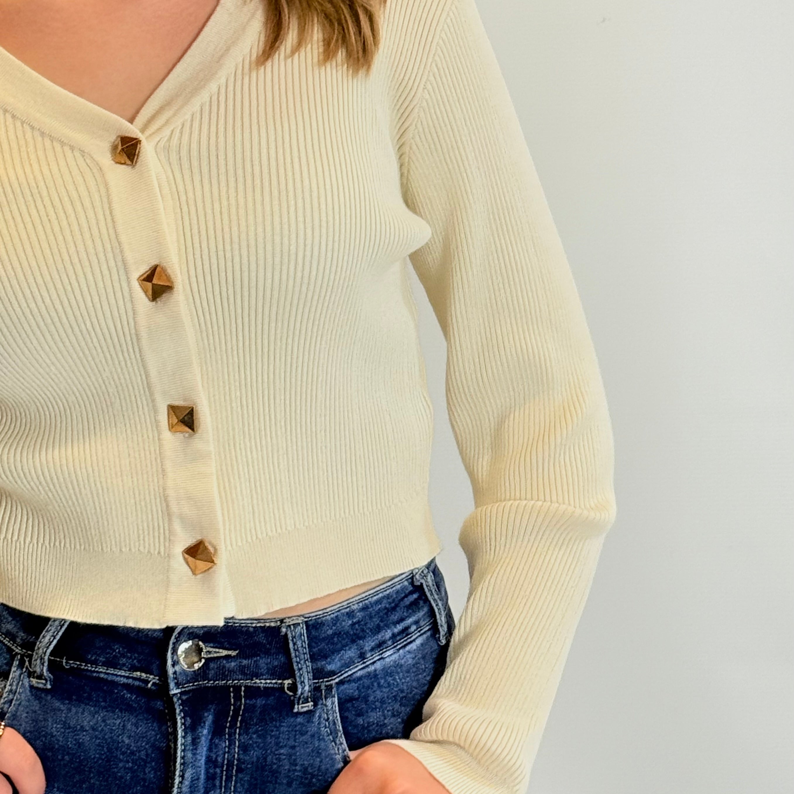 Person wearing a cream ribbed cardigan with gold buttons and blue jeans on a light gray background