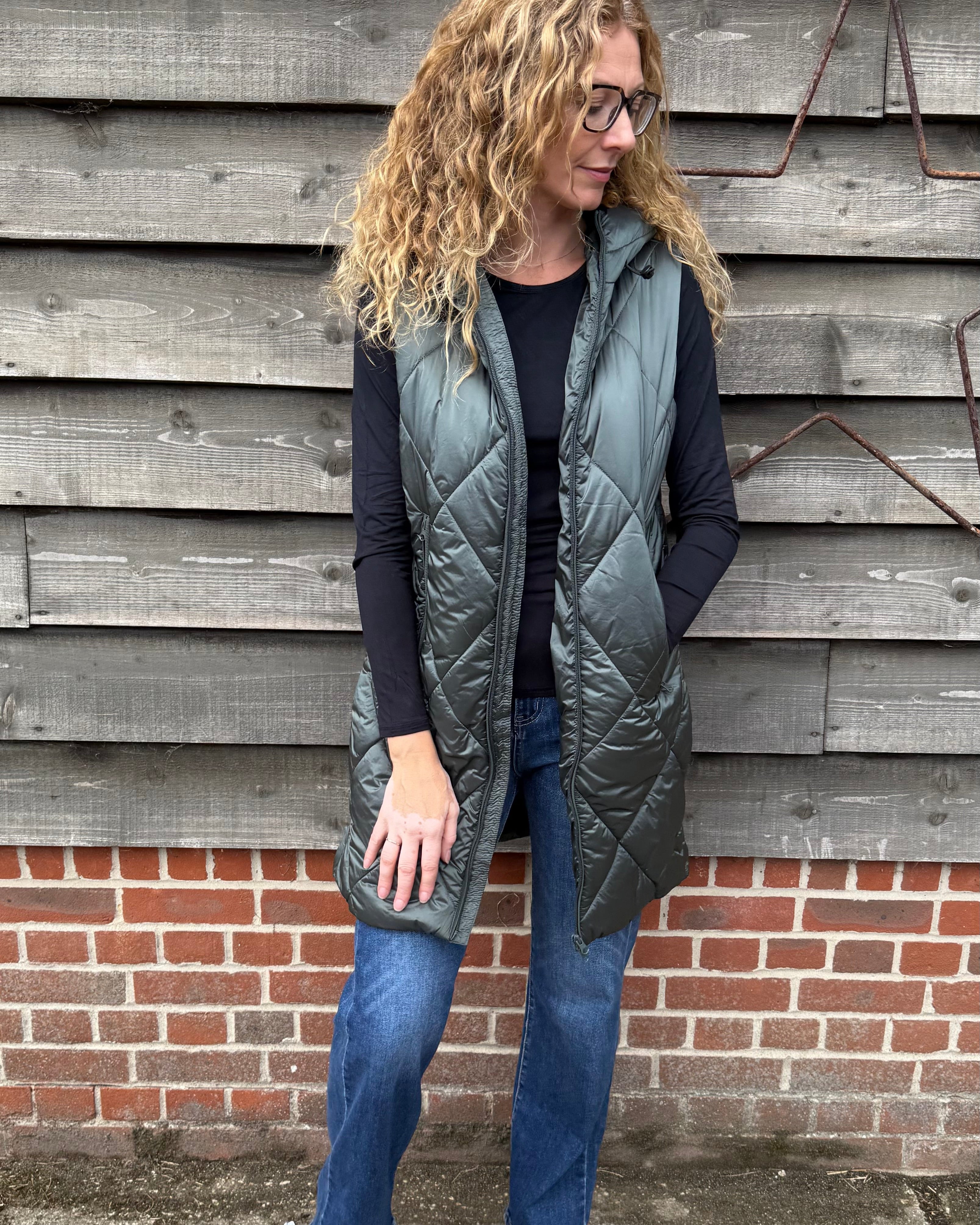Woman wearing a long gray quilted vest standing against a wooden wall.