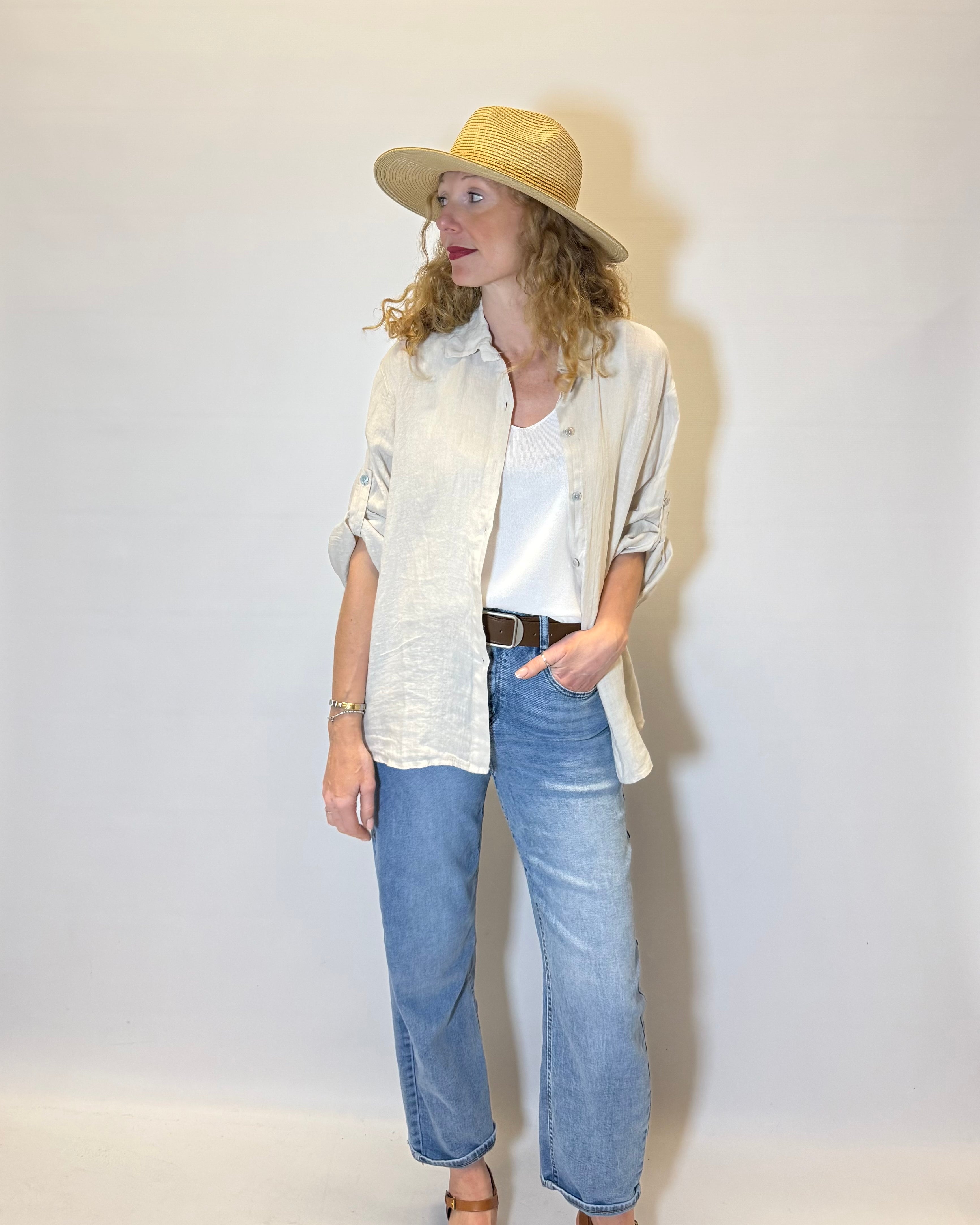 Person wearing a beige hat, light-colored shirt, white top, blue jeans, and brown sandals against a white background