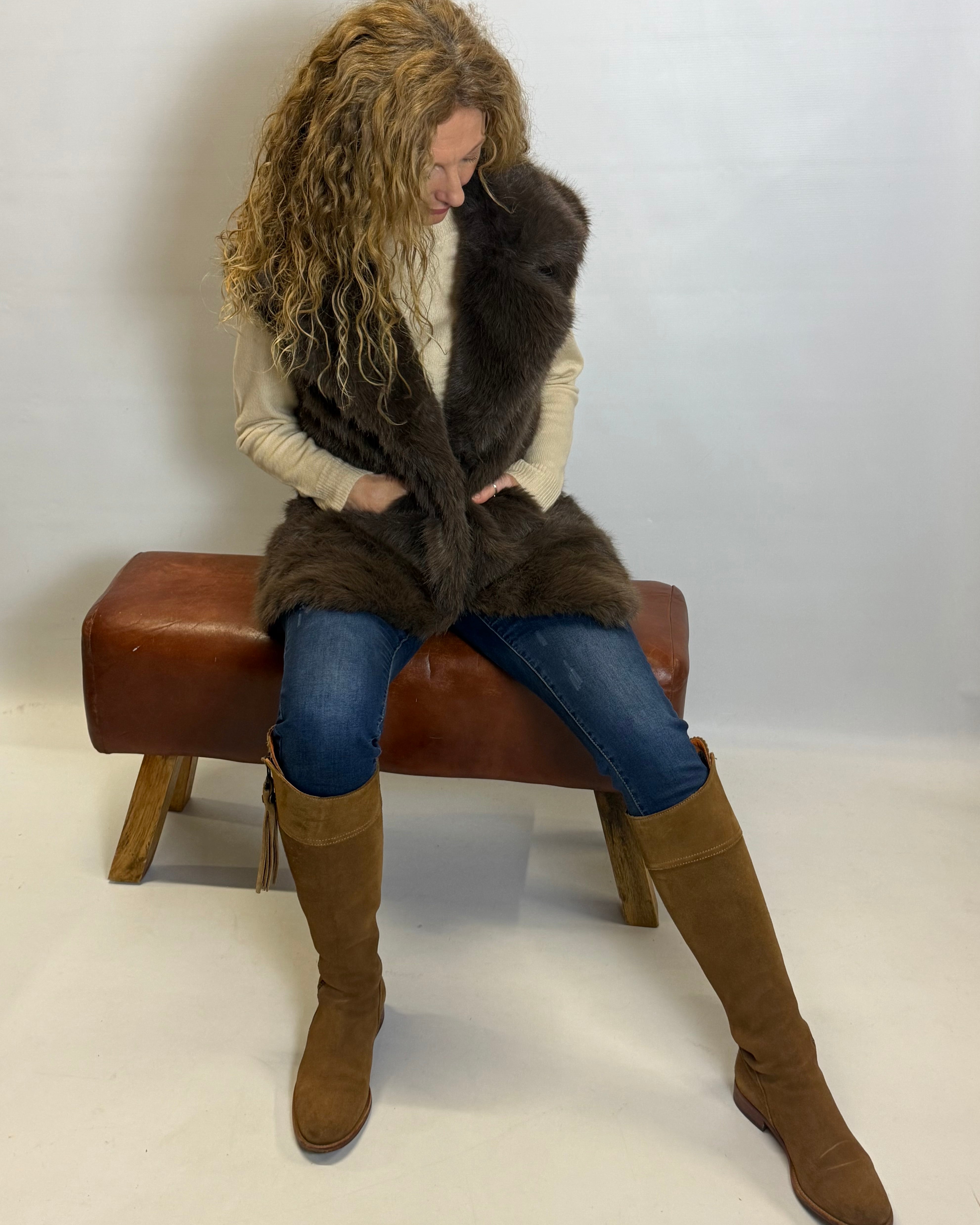 Woman sitting on a brown bench wearing a fur vest and knee-high boots against a white background