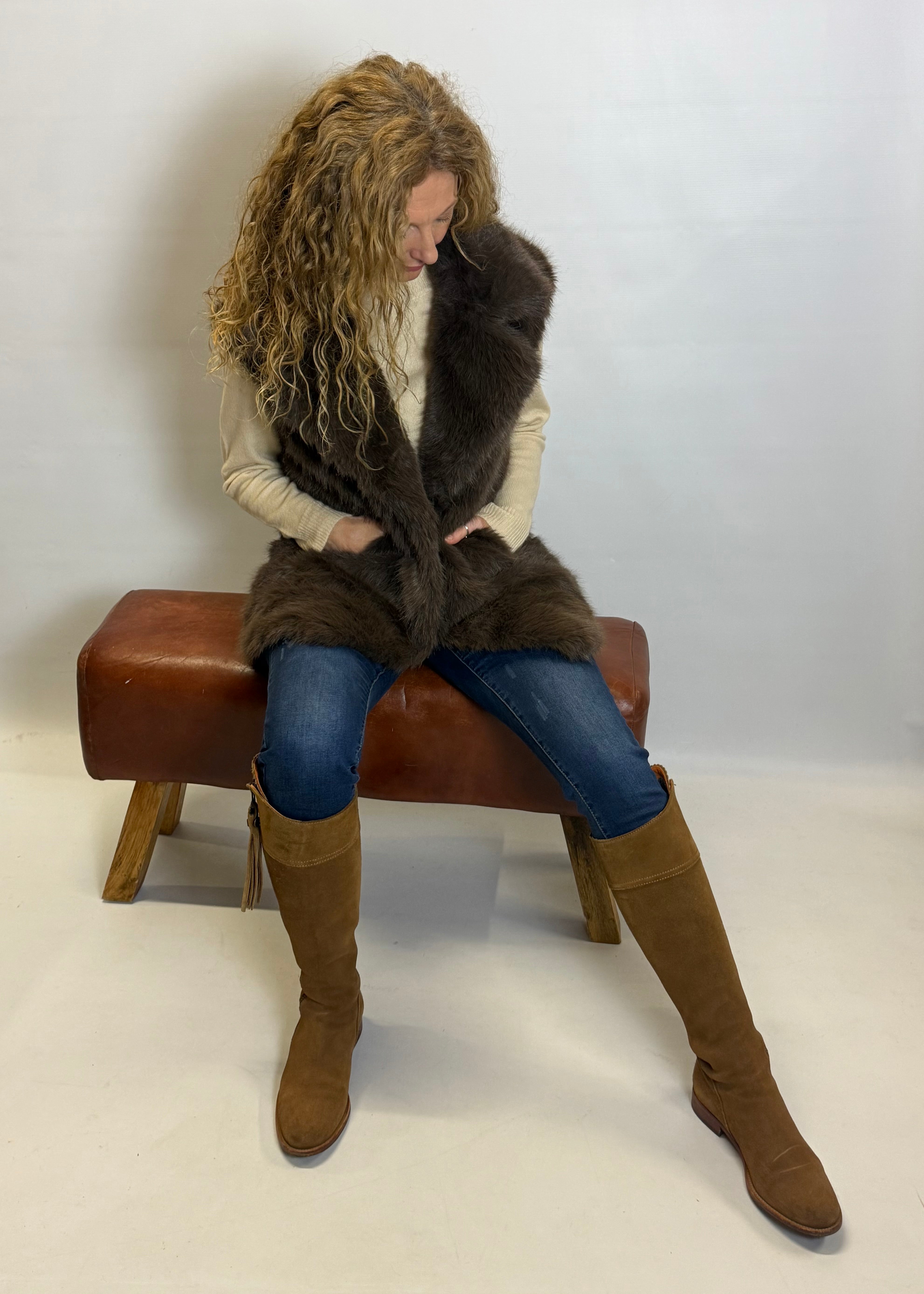 Woman sitting on a brown bench wearing a fur vest and knee-high boots against a white background
