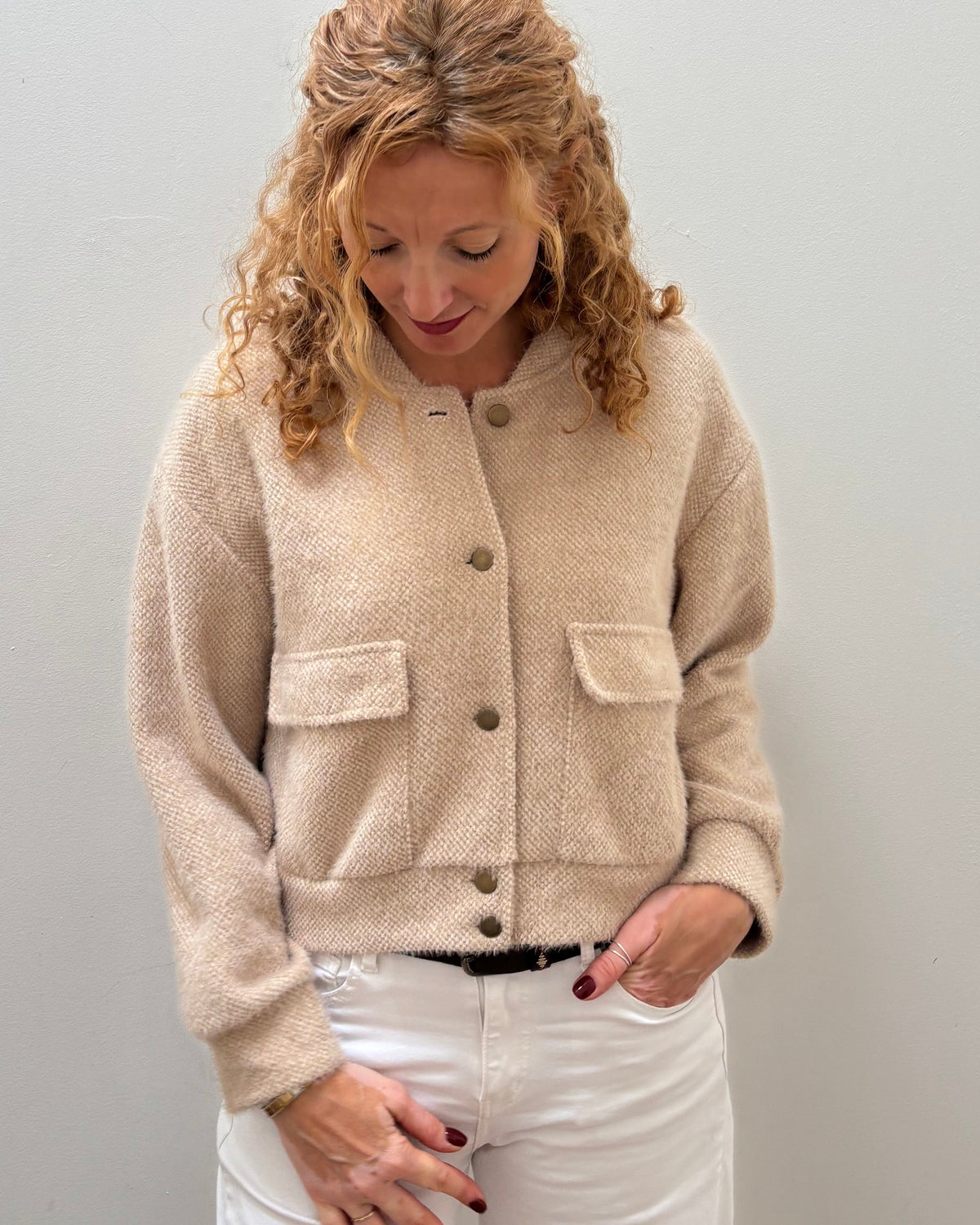 A woman with blonde curly hair wearing a beige fuzzy cropped cardigan with button details and white pants, standing against a plain light gray wall.