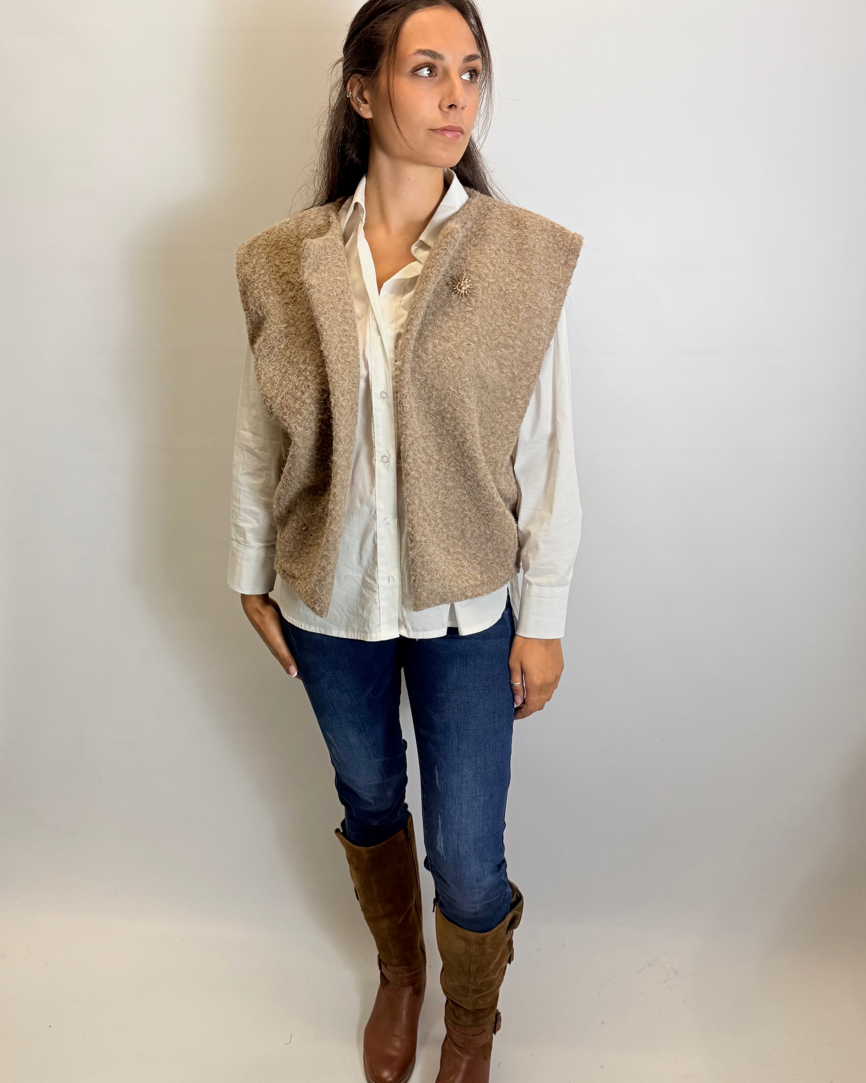Woman wearing a beige vest over a white shirt, blue jeans, and brown boots on a plain background