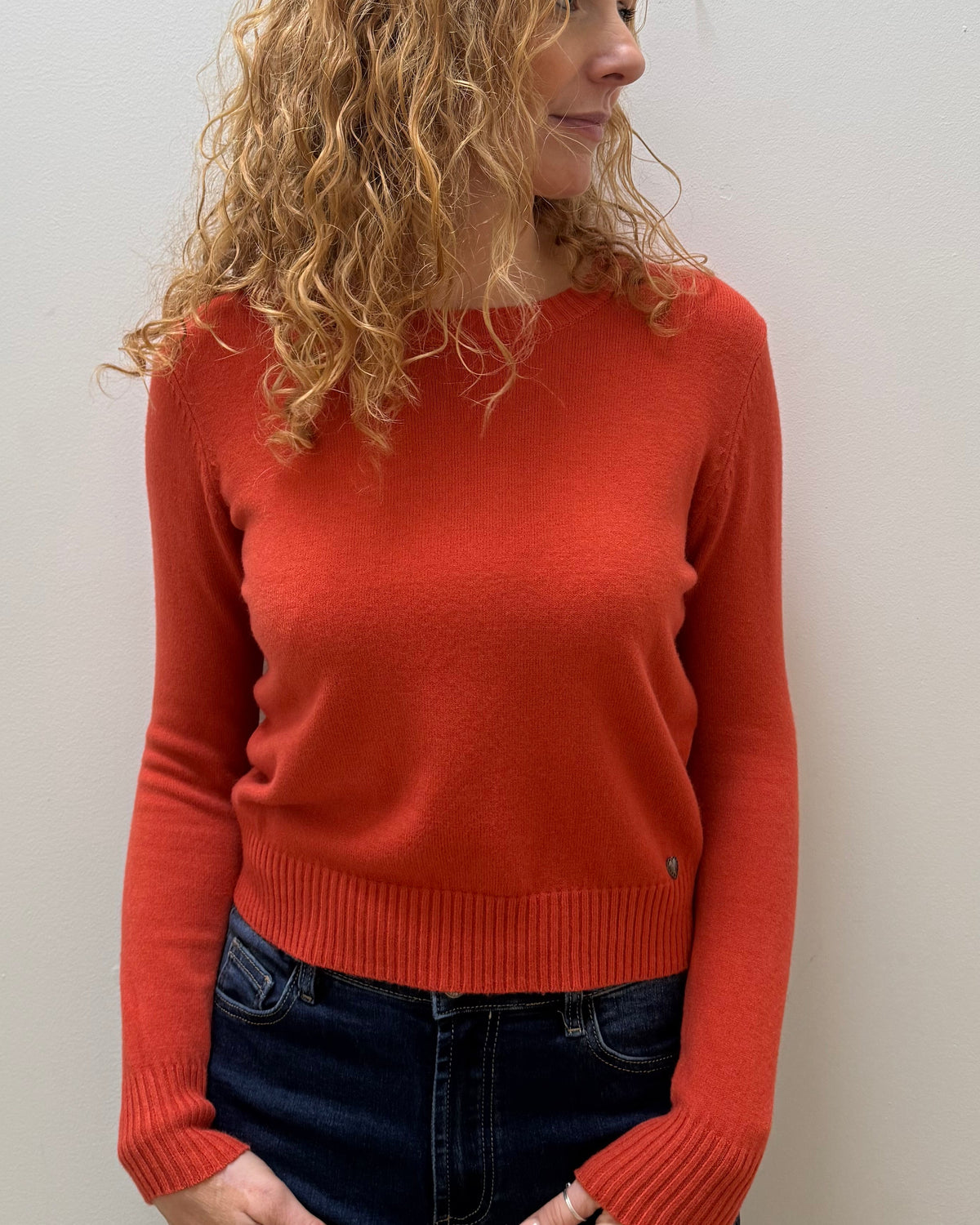 A woman with curly blonde hair wearing a rust-orange long-sleeve knit sweater and dark blue jeans, standing against a plain off-white wall.