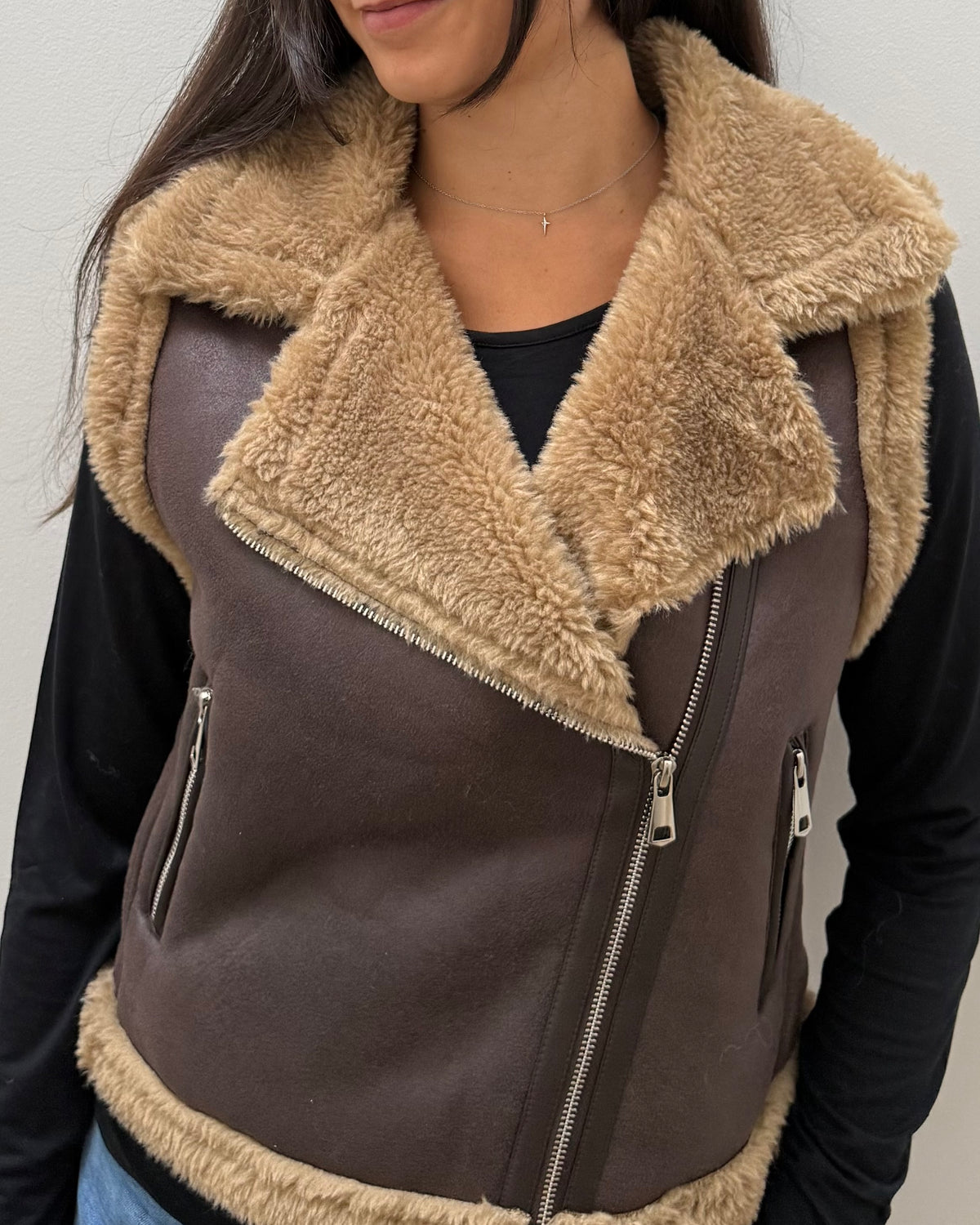 A close-up of a person wearing a brown faux-leather aviator-style vest with a thick beige sherpa collar and lining, over a black long-sleeve top.