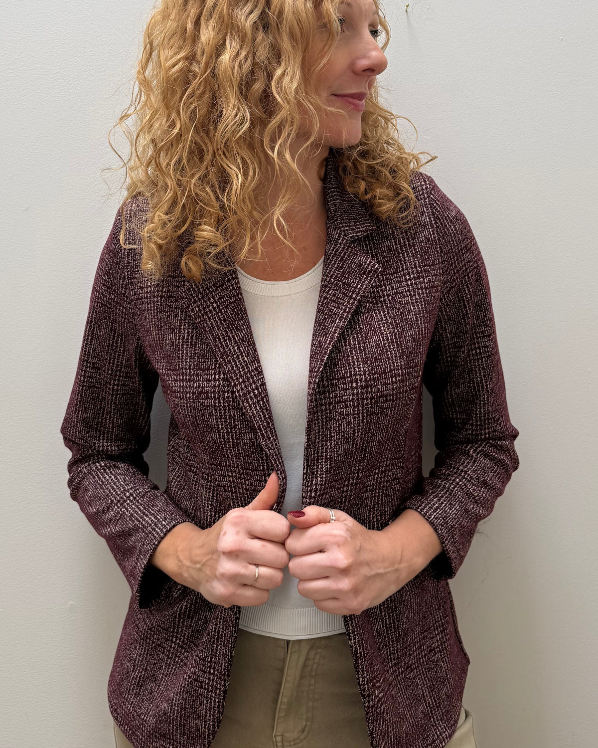 A woman with curly blonde hair wearing a textured plum purple blazer over a white top and khaki pants, standing against a plain light gray wall.