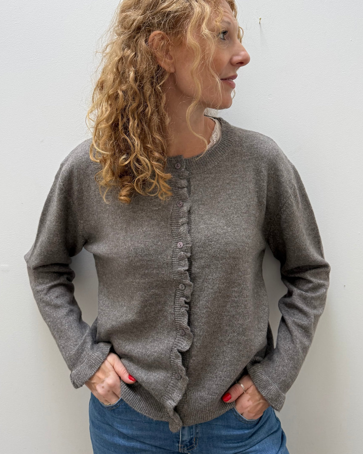 A woman with curly blonde hair wearing a gray knit cardigan sweater with ruffle trim down the front and blue jeans, standing against a plain light gray wall.