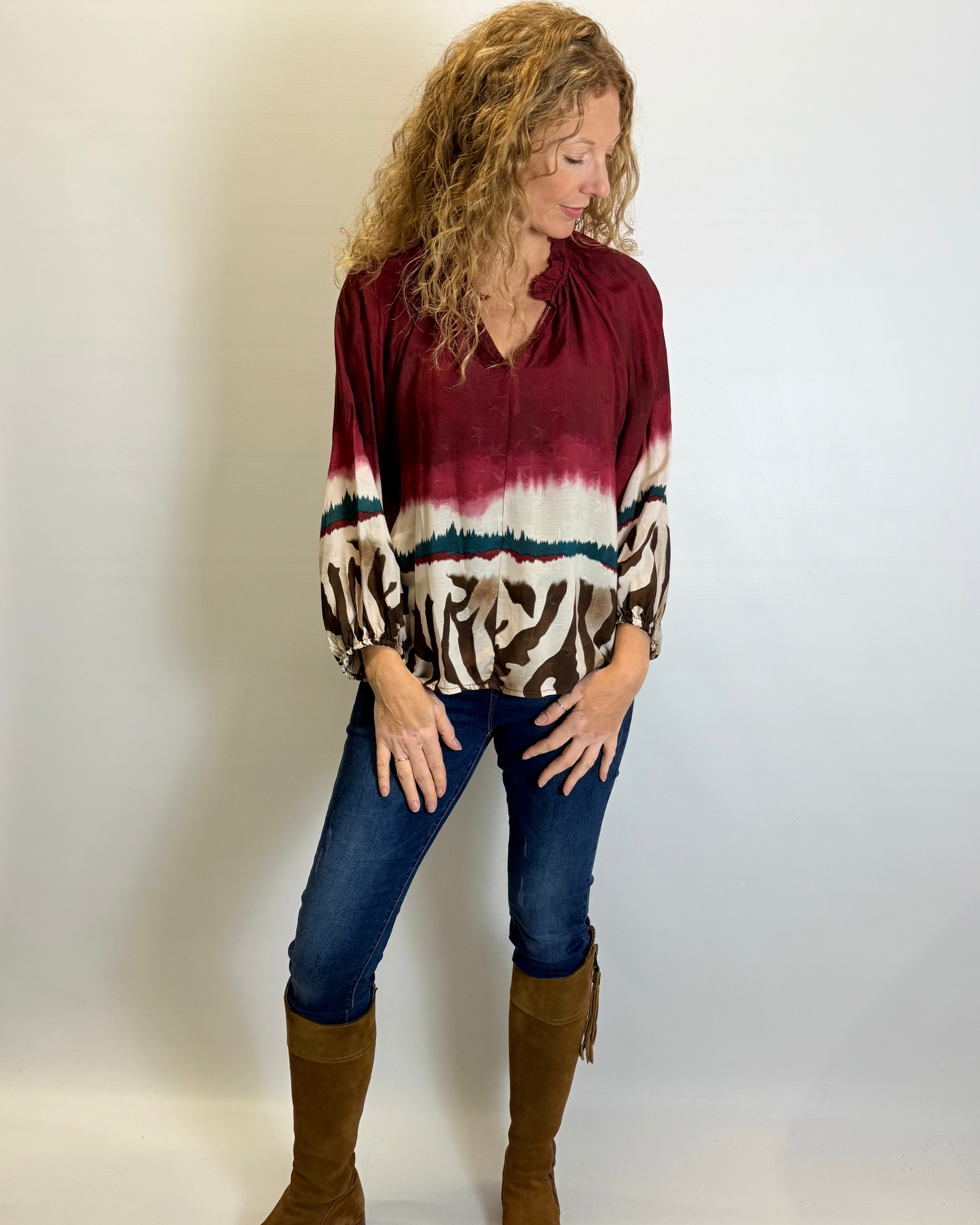 Woman wearing a maroon tie-dye blouse with animal print sleeves, blue jeans, and brown boots against a white background.