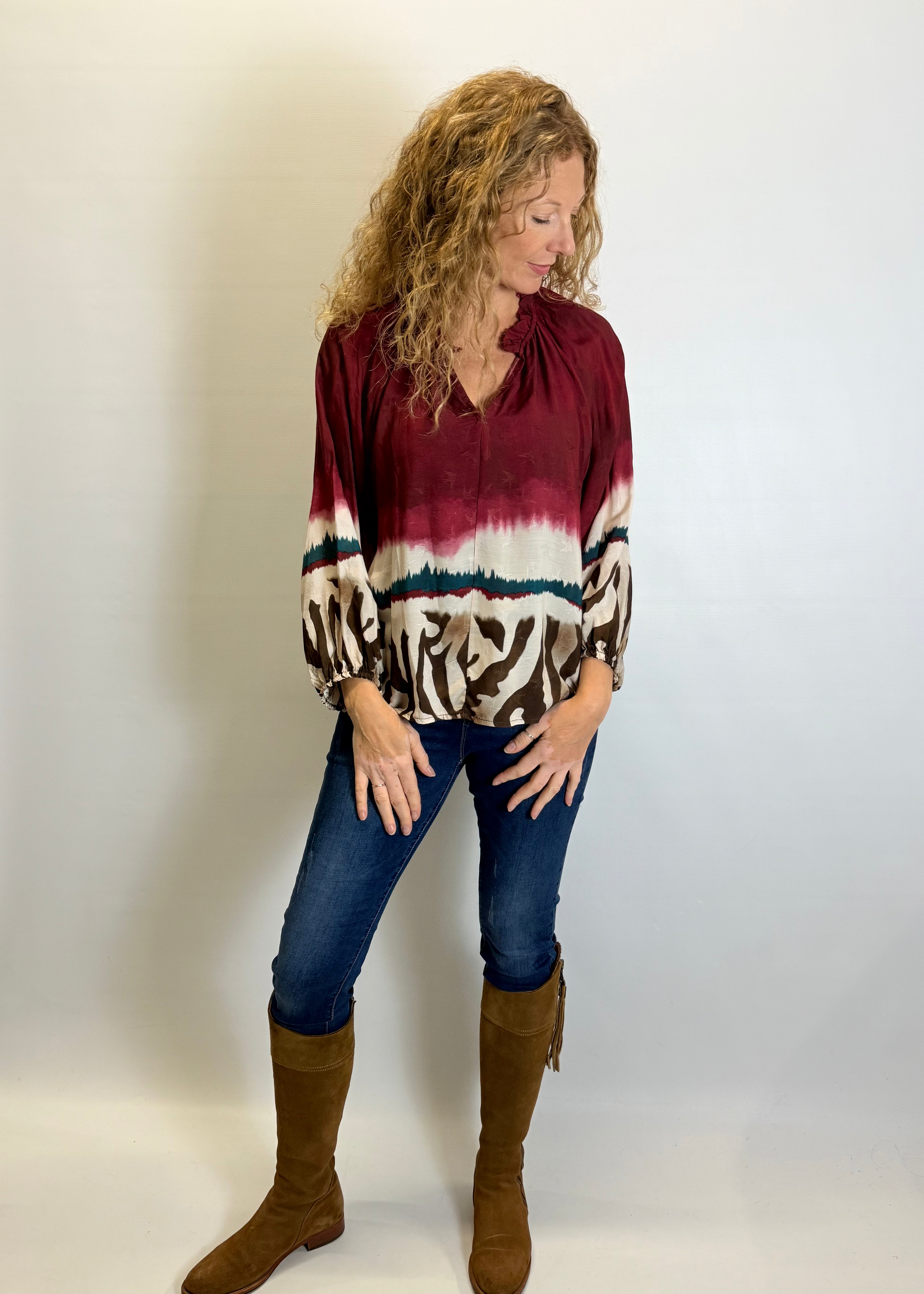 Woman wearing a maroon tie-dye blouse with animal print sleeves, blue jeans, and brown boots against a white background.