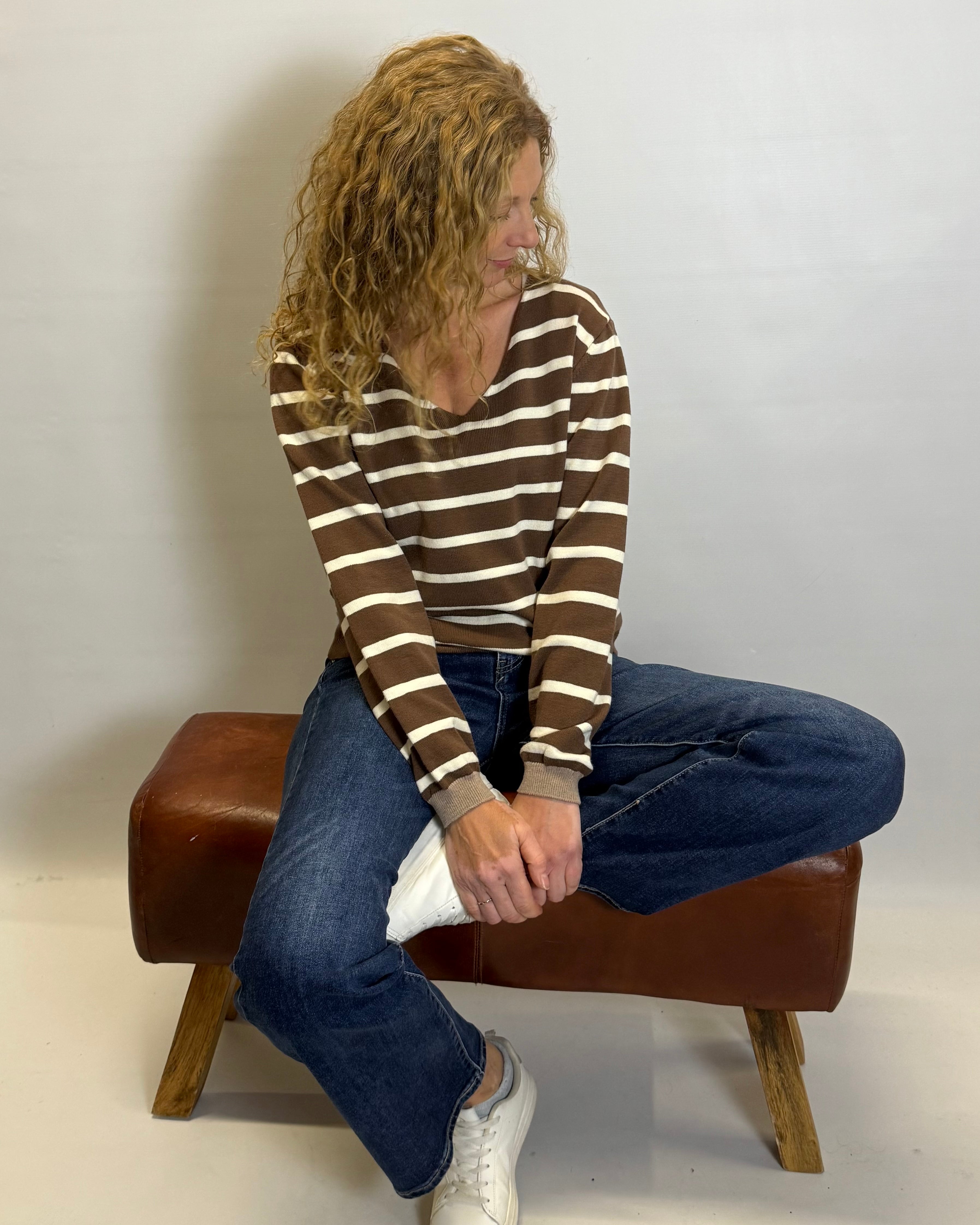 Person sitting on a brown stool wearing a striped sweater and jeans against a white background