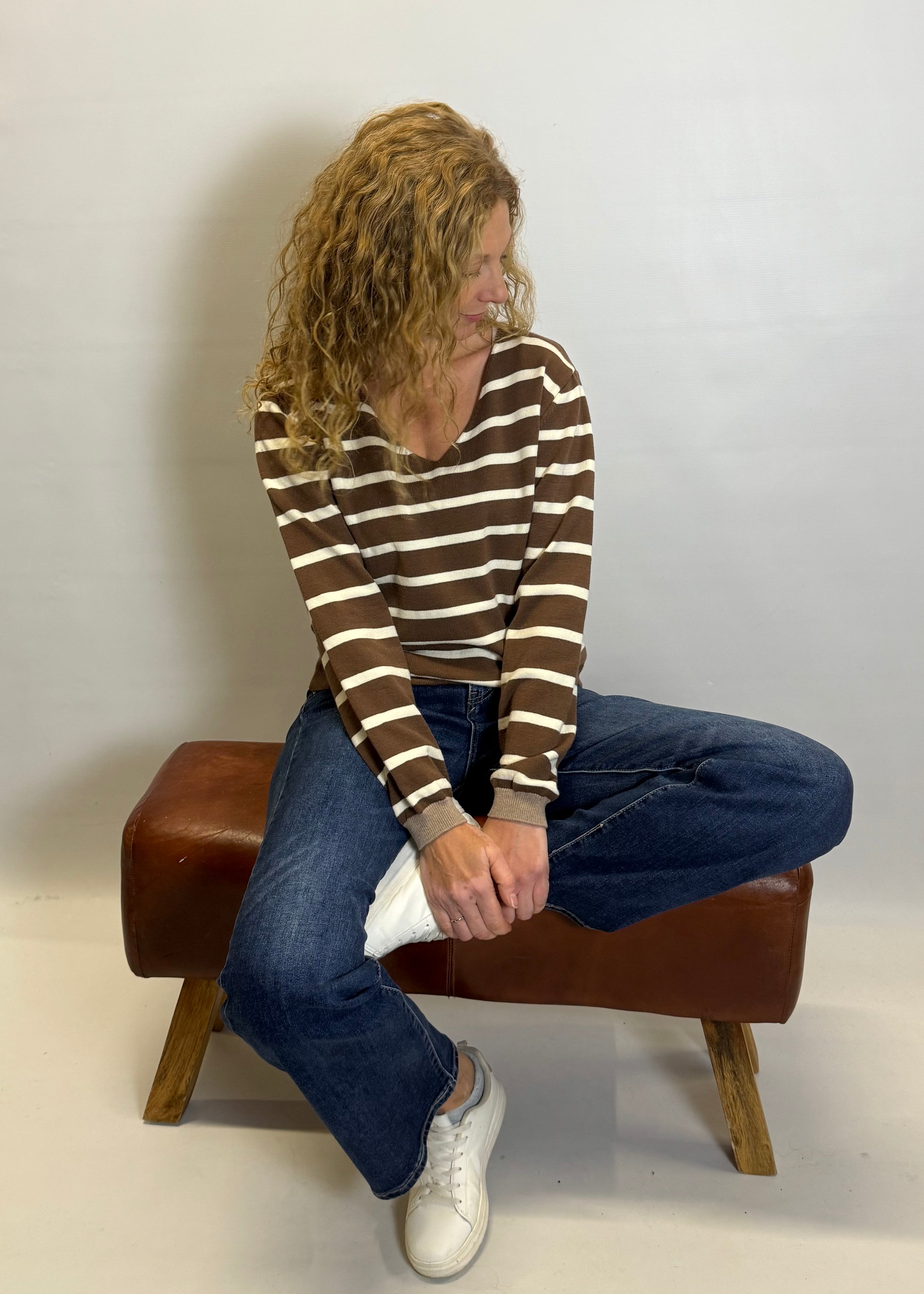 Person sitting on a brown stool wearing a striped sweater and jeans against a white background