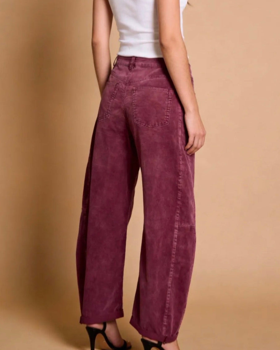 A model from the back wearing high-waisted, wide-leg corduroy pants in a deep burgundy or plum shade, paired with a white tank top and black high-heeled sandals, against a solid beige studio backdrop.