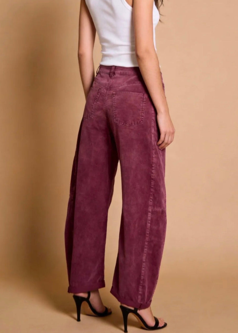A model from the back wearing high-waisted, wide-leg corduroy pants in a deep burgundy or plum shade, paired with a white tank top and black high-heeled sandals, against a solid beige studio backdrop.
