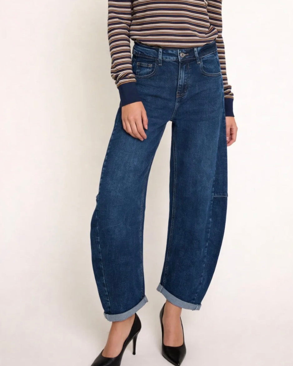 A model wearing dark blue wide-leg cuffed denim jeans, paired with a brown and beige striped long-sleeve top and black pointed-toe heels, standing against a plain off-white background.