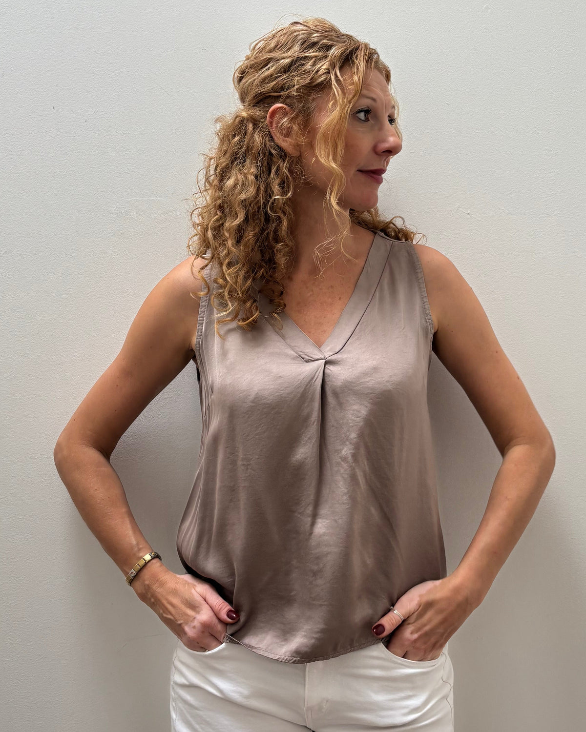 A woman with curly blonde hair stands against a plain light gray wall wearing a sleeveless taupe satin-like top and white pants.