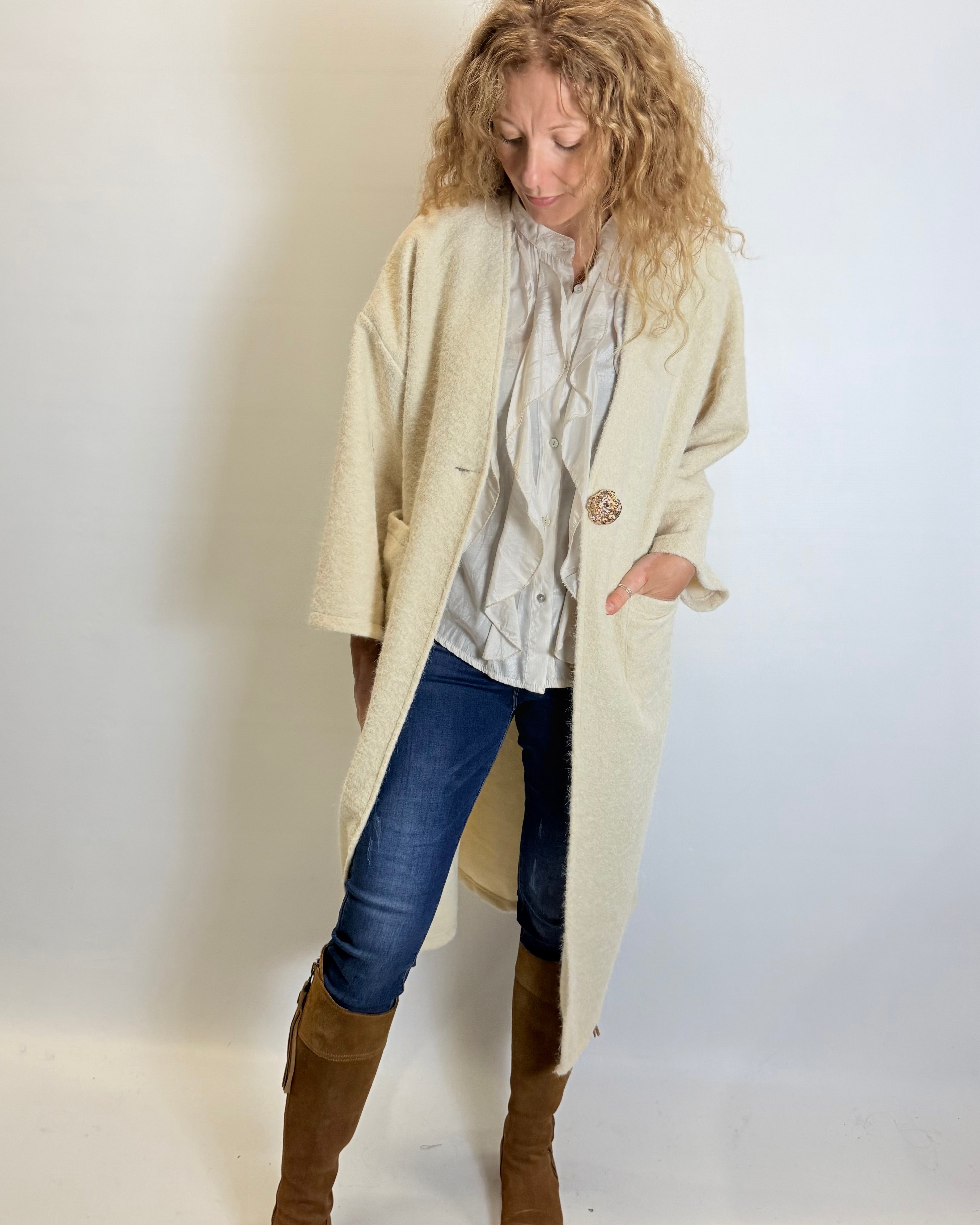 Woman wearing a beige coat, blue jeans, and brown boots against a plain background