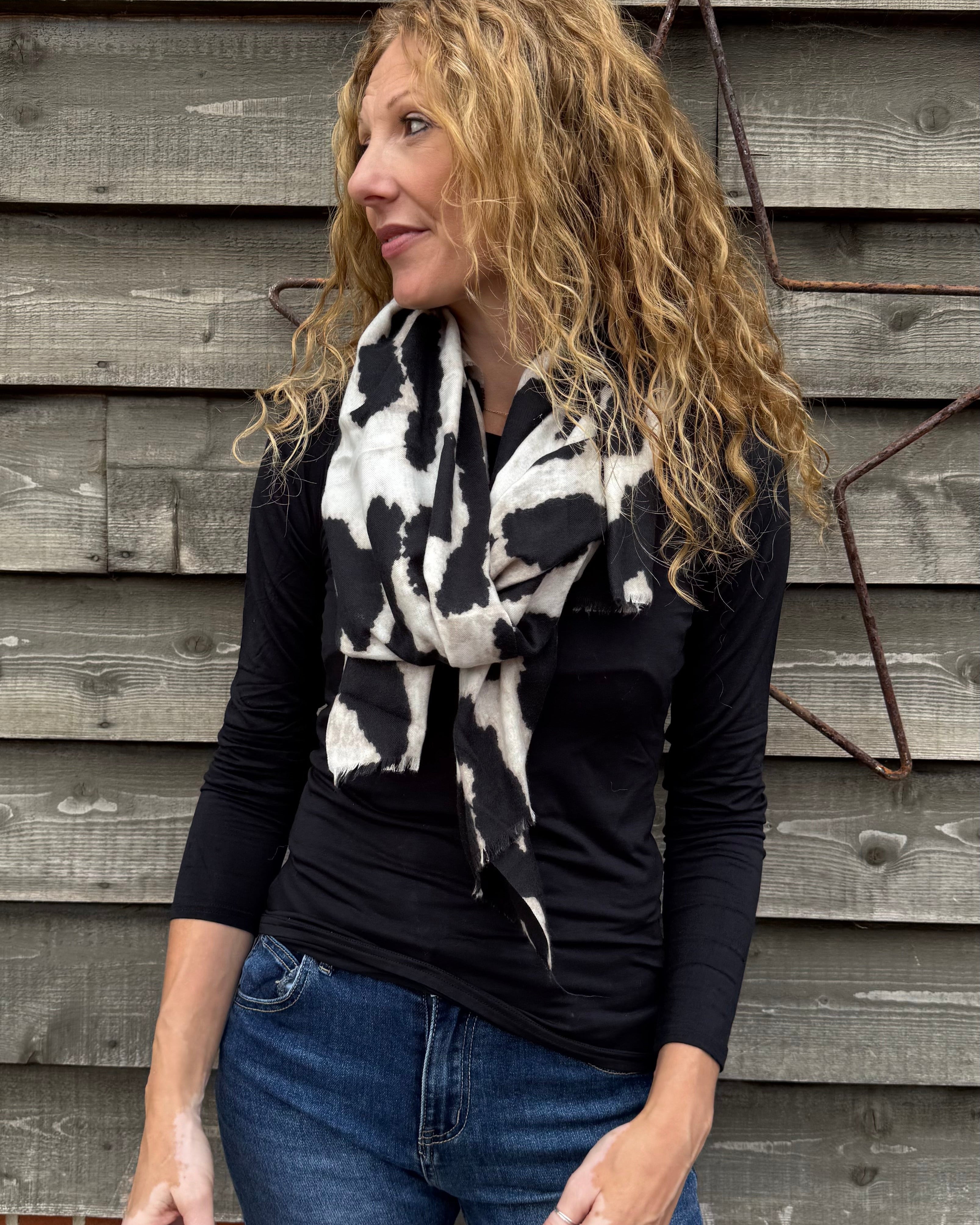 Woman wearing a black top and blue jeans with a black and white patterned scarf against a wooden wall.