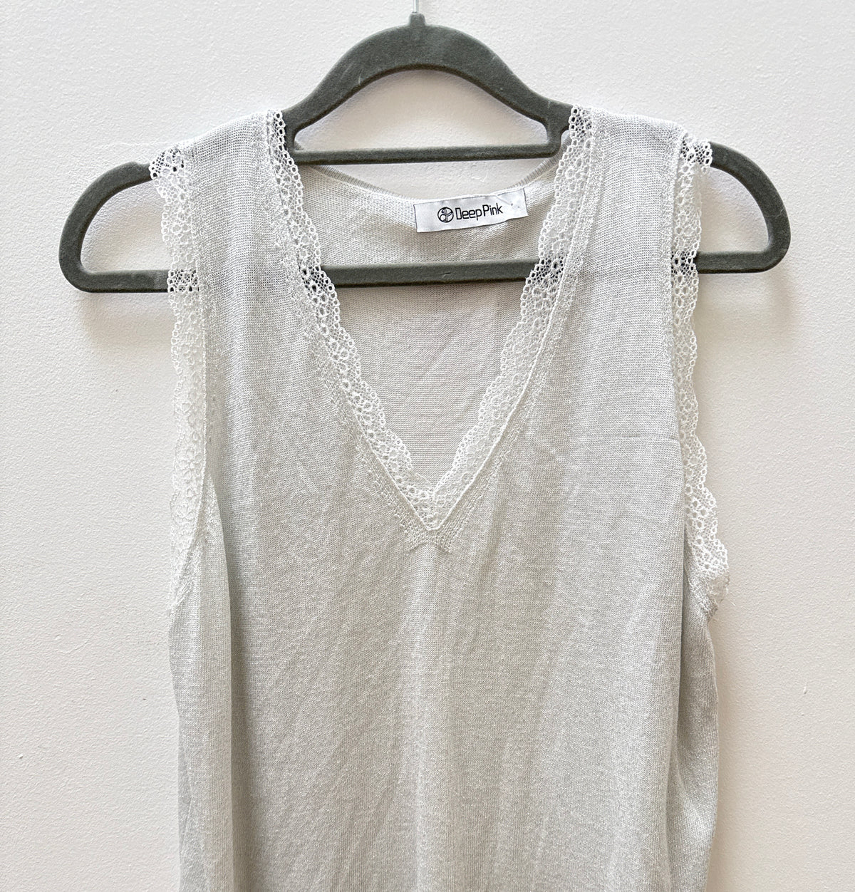 A light gray sleeveless V-neck top with delicate lace trim along the armholes and neckline, hanging on a black hanger against a plain off-white wall.