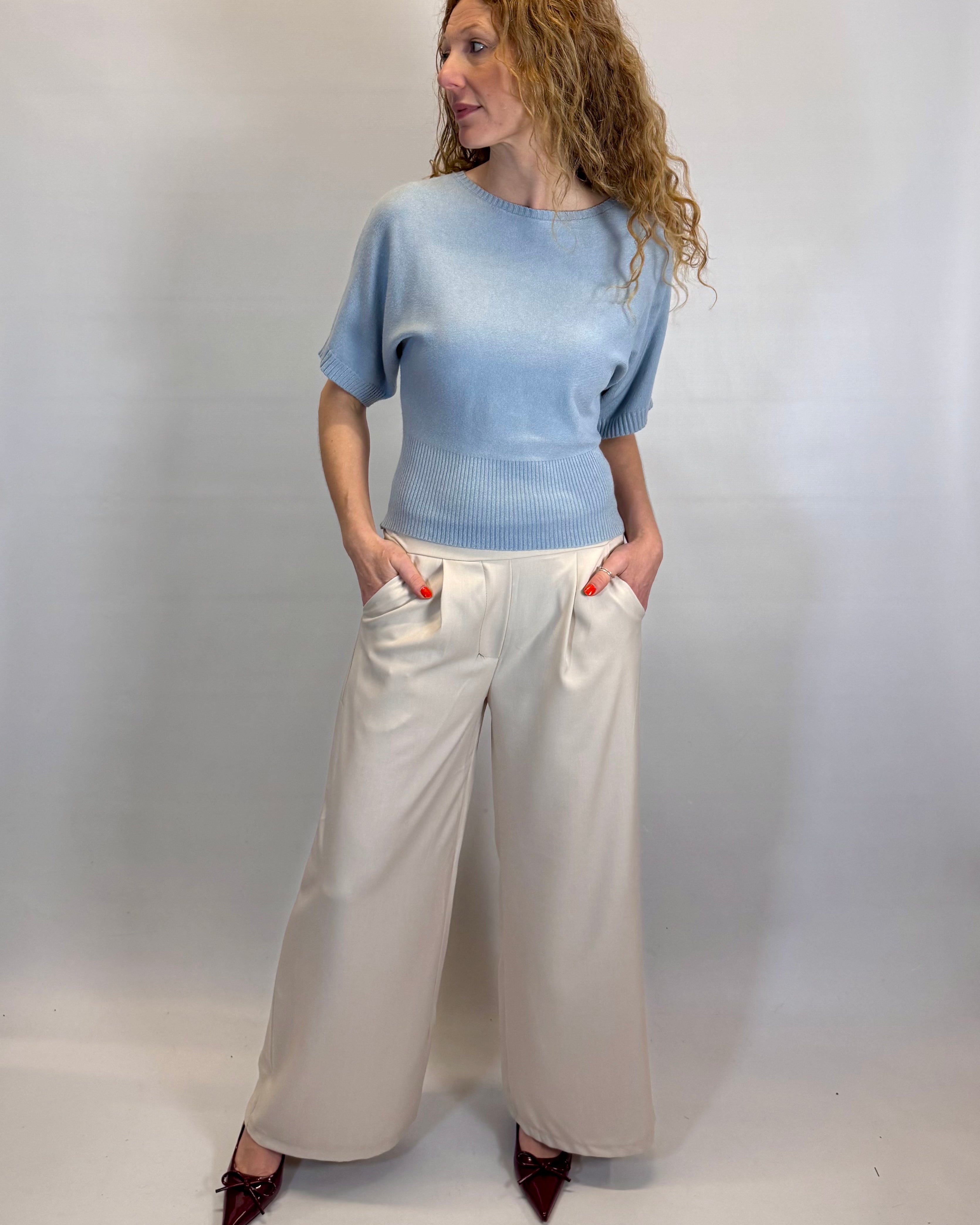Woman wearing a light blue sweater and white pants against a plain background