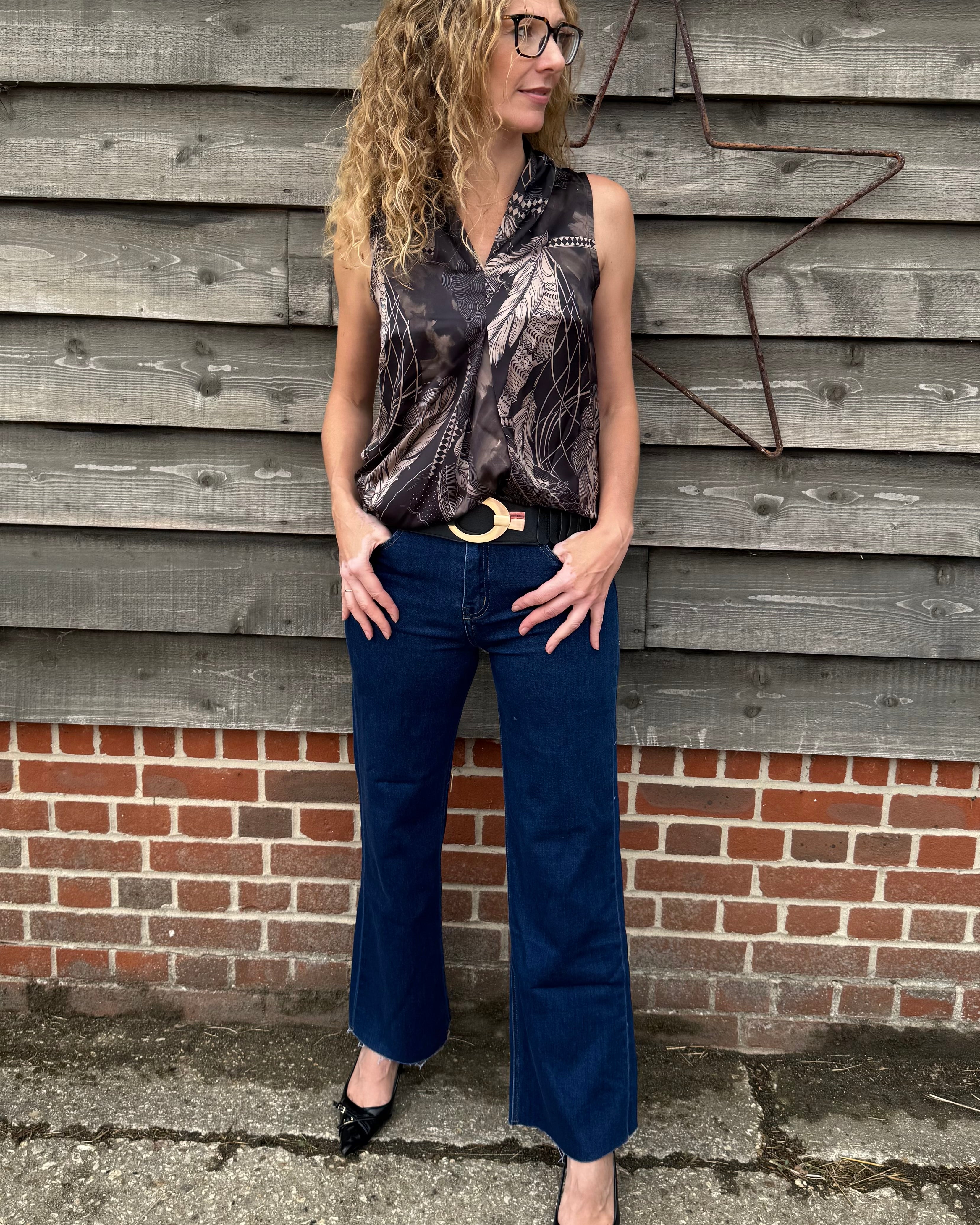 Woman wearing a patterned top and blue jeans standing against a wooden wall.
