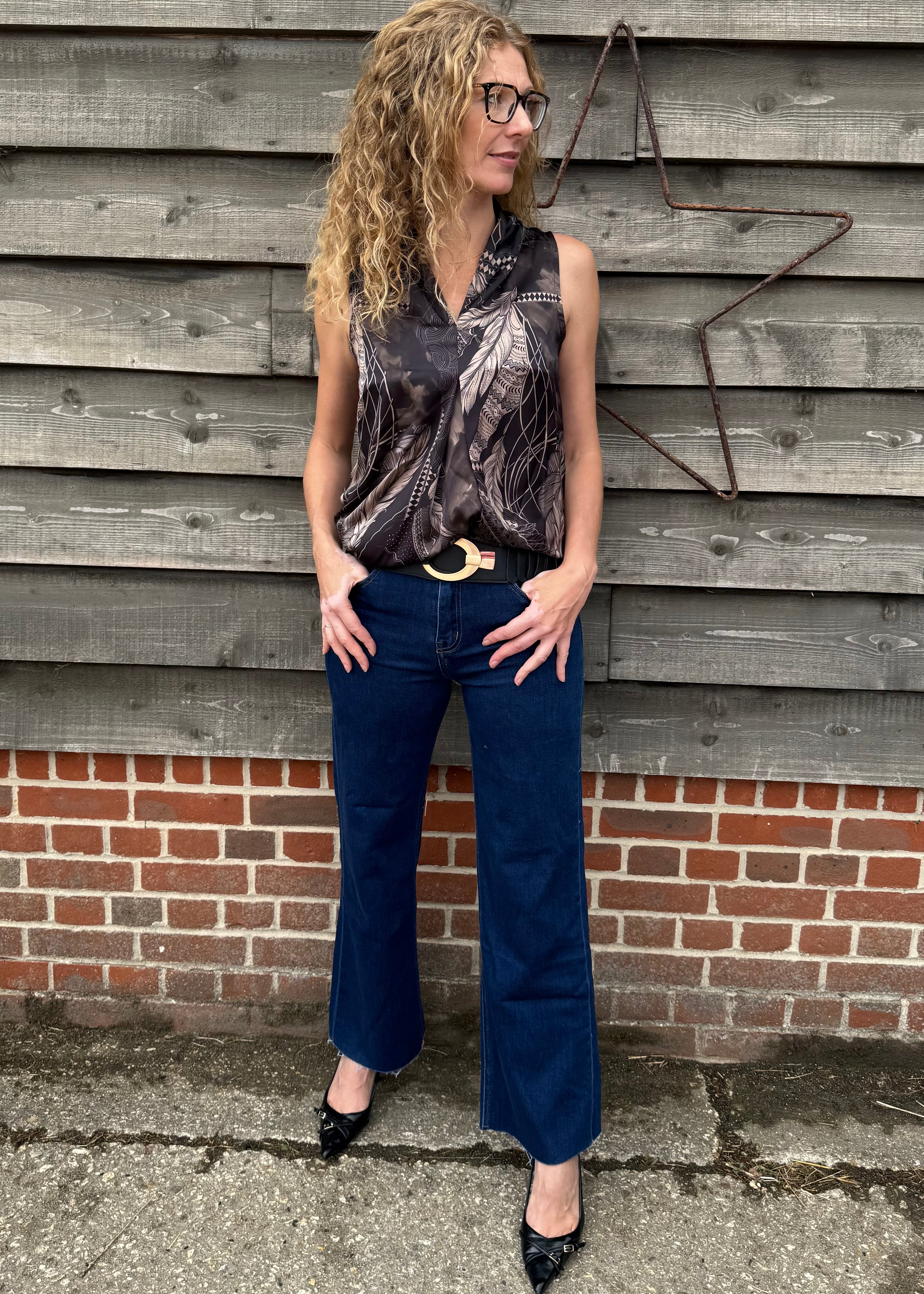 Woman wearing a patterned top and blue jeans standing against a wooden wall.