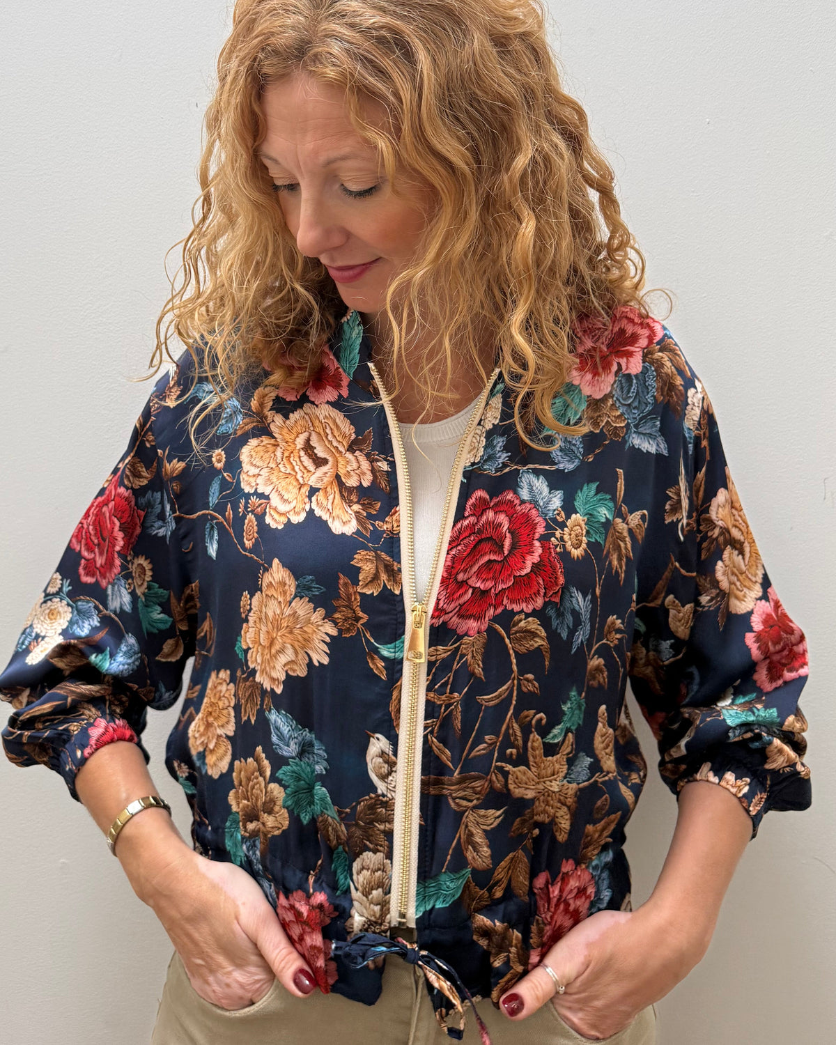A woman with curly blonde hair wearing a navy blue floral bomber jacket with red, gold, and green flowers, paired with beige pants, standing against a plain light gray wall.