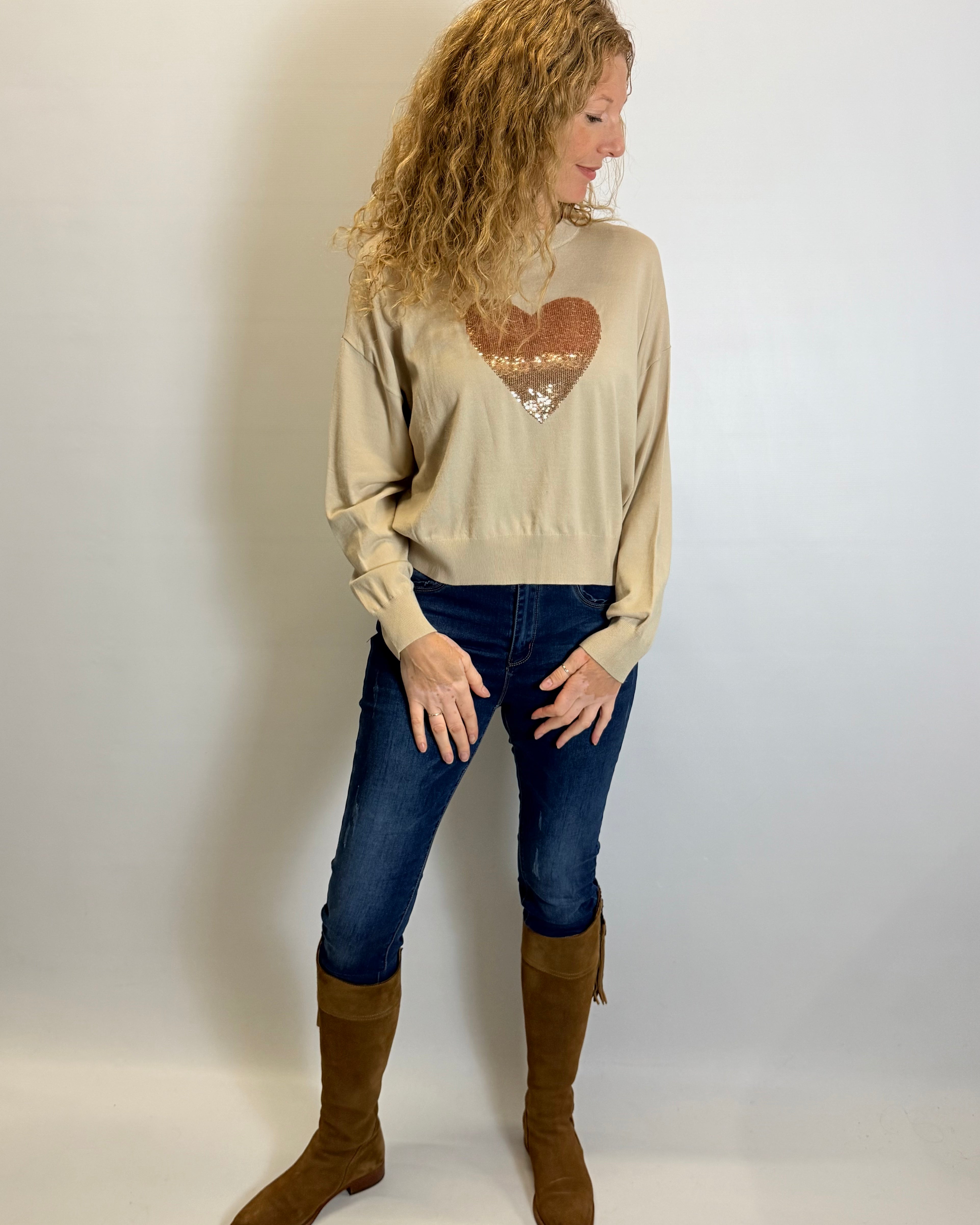 Woman wearing a beige sweater with a heart design, blue jeans, and brown boots against a white background