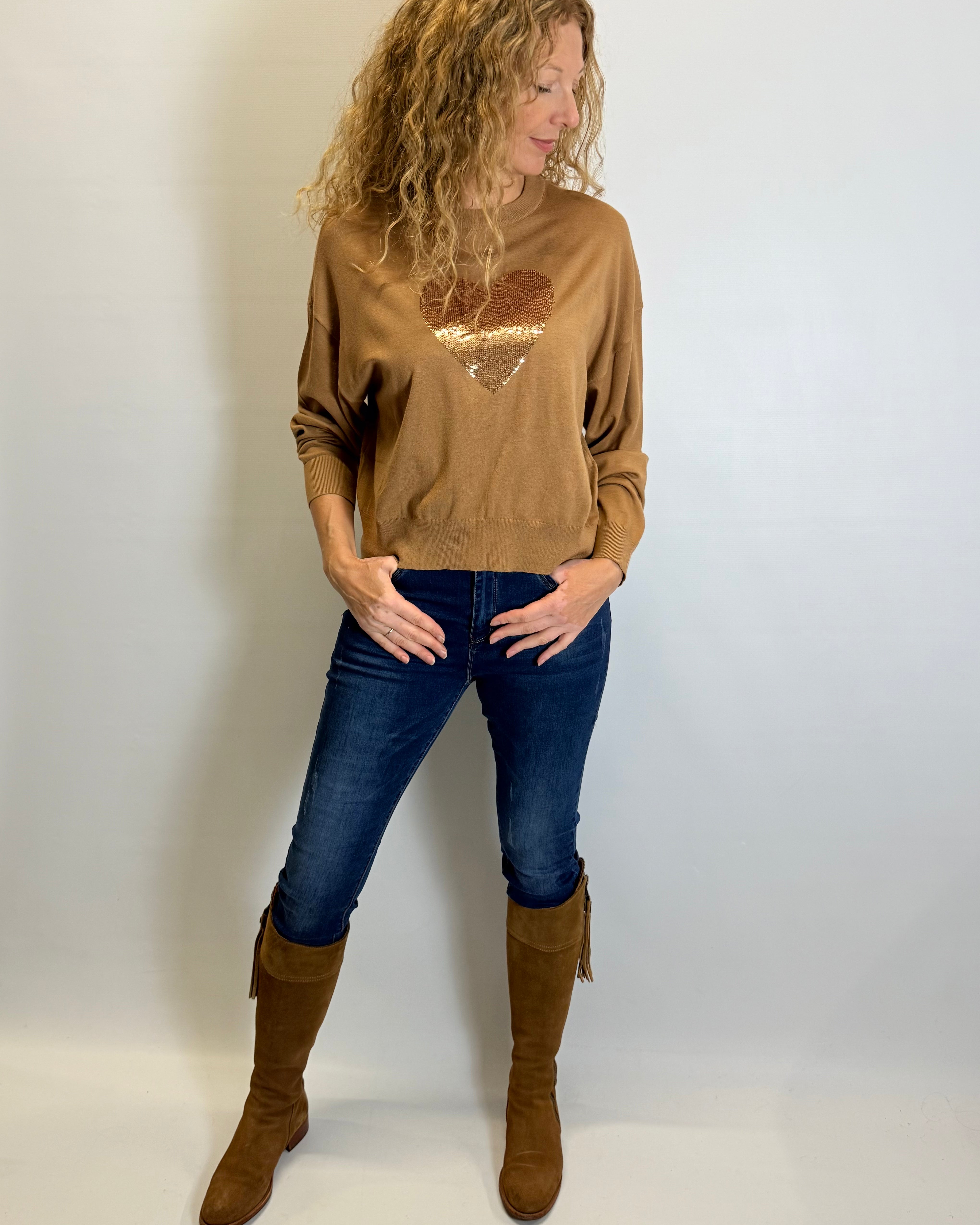 Woman wearing a brown sweater with a heart design, blue jeans, and brown knee-high boots against a white background.
