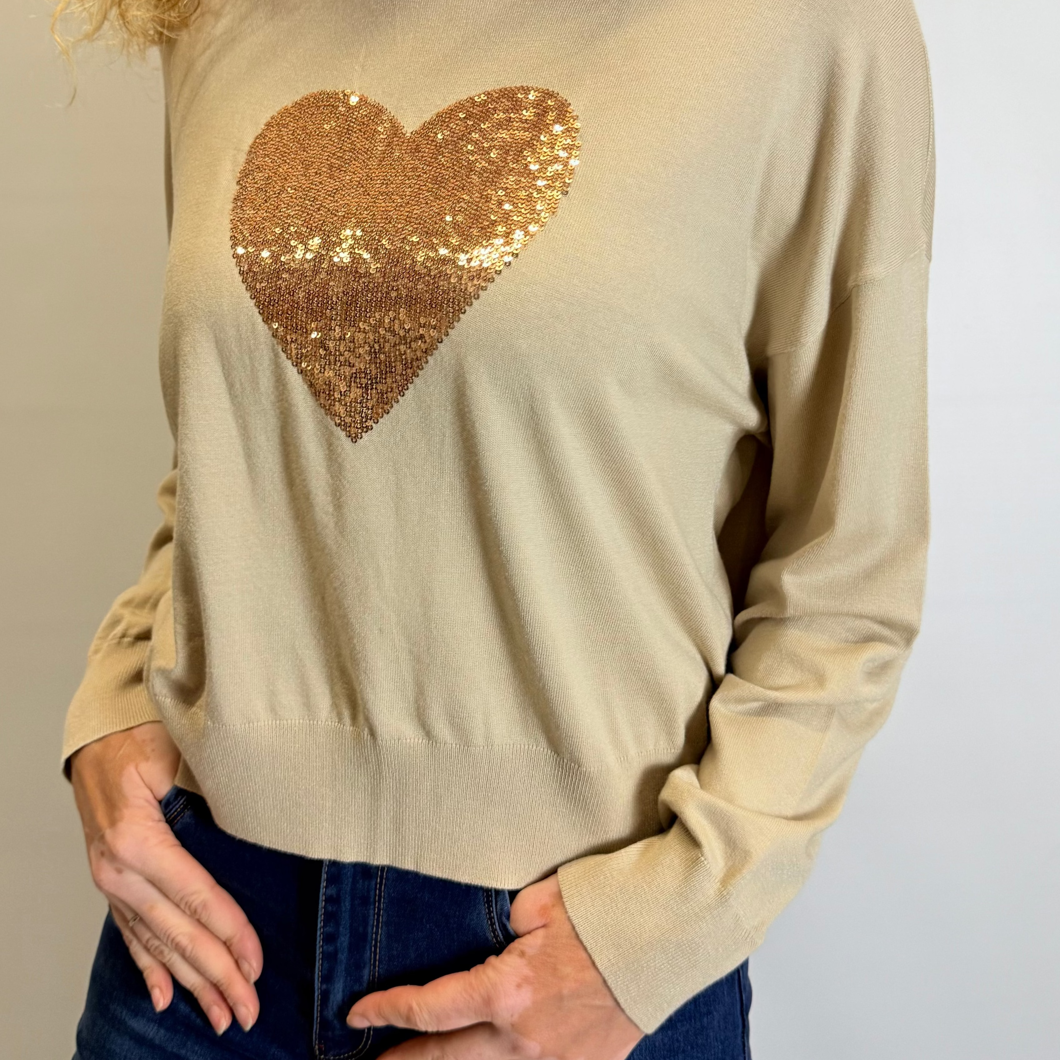 Woman wearing a beige sweater with a gold sequin heart design against a plain background