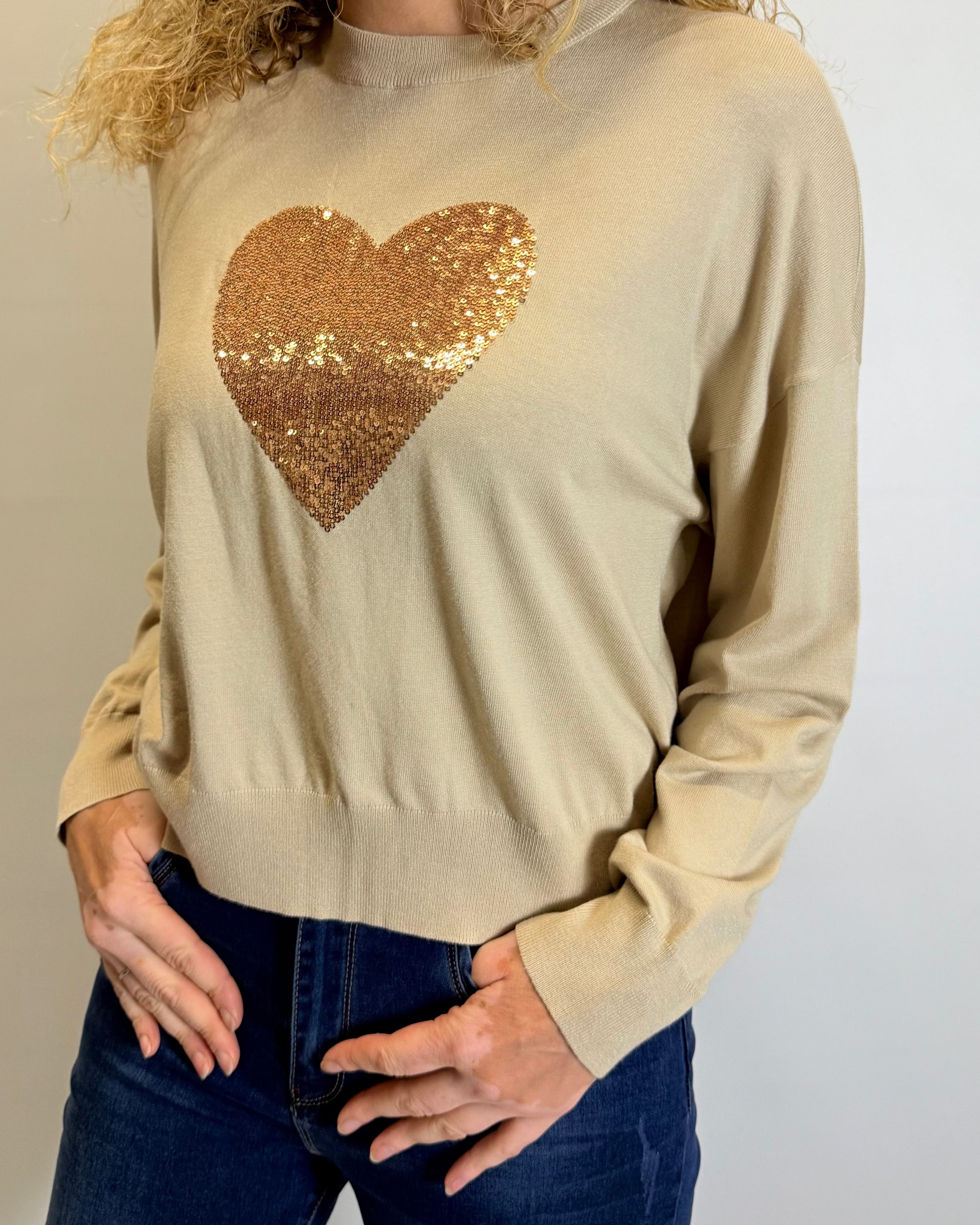 Woman wearing a beige sweater with a gold sequin heart design against a plain background