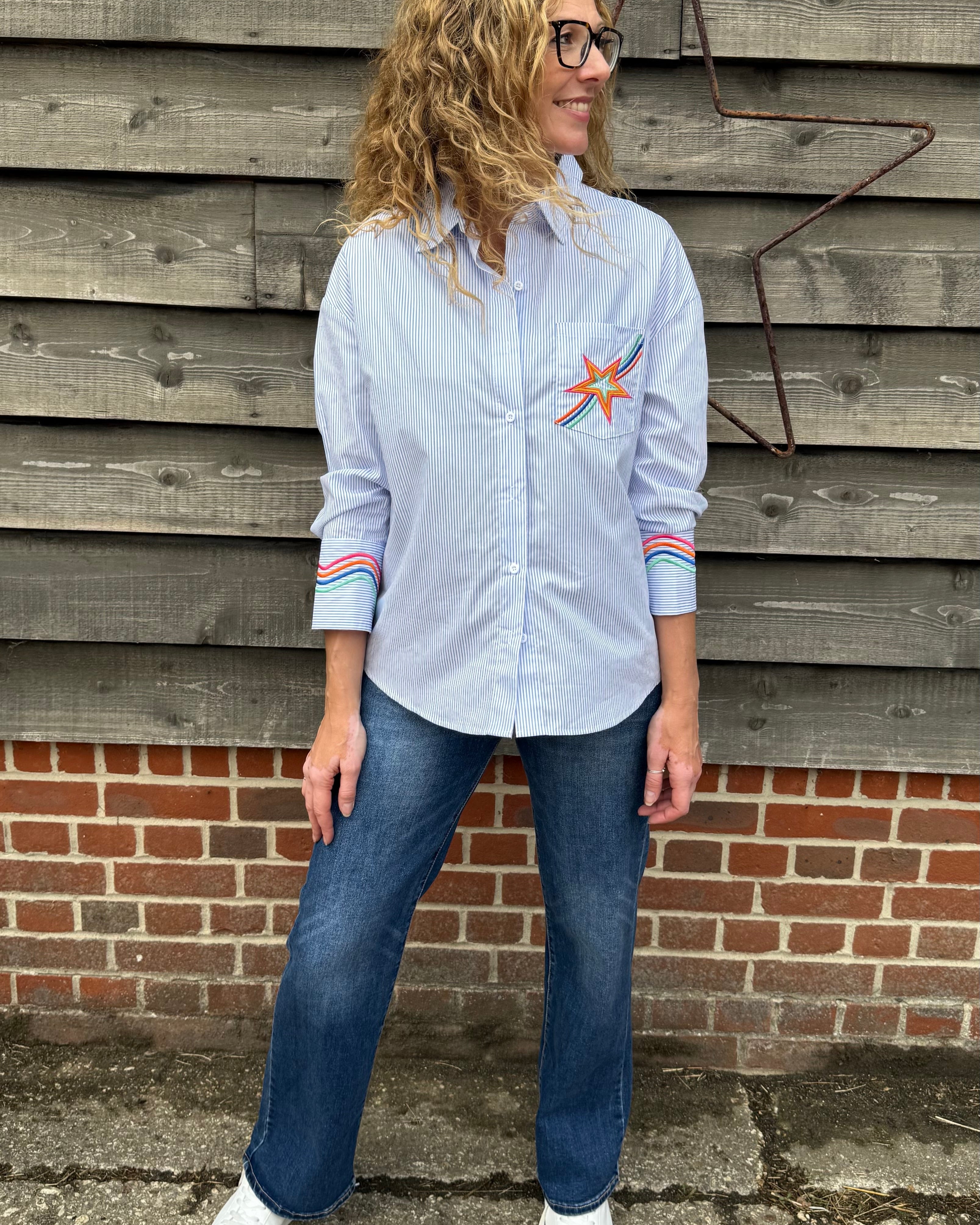 Person wearing a light blue shirt with a star design and jeans, standing against a wooden wall.