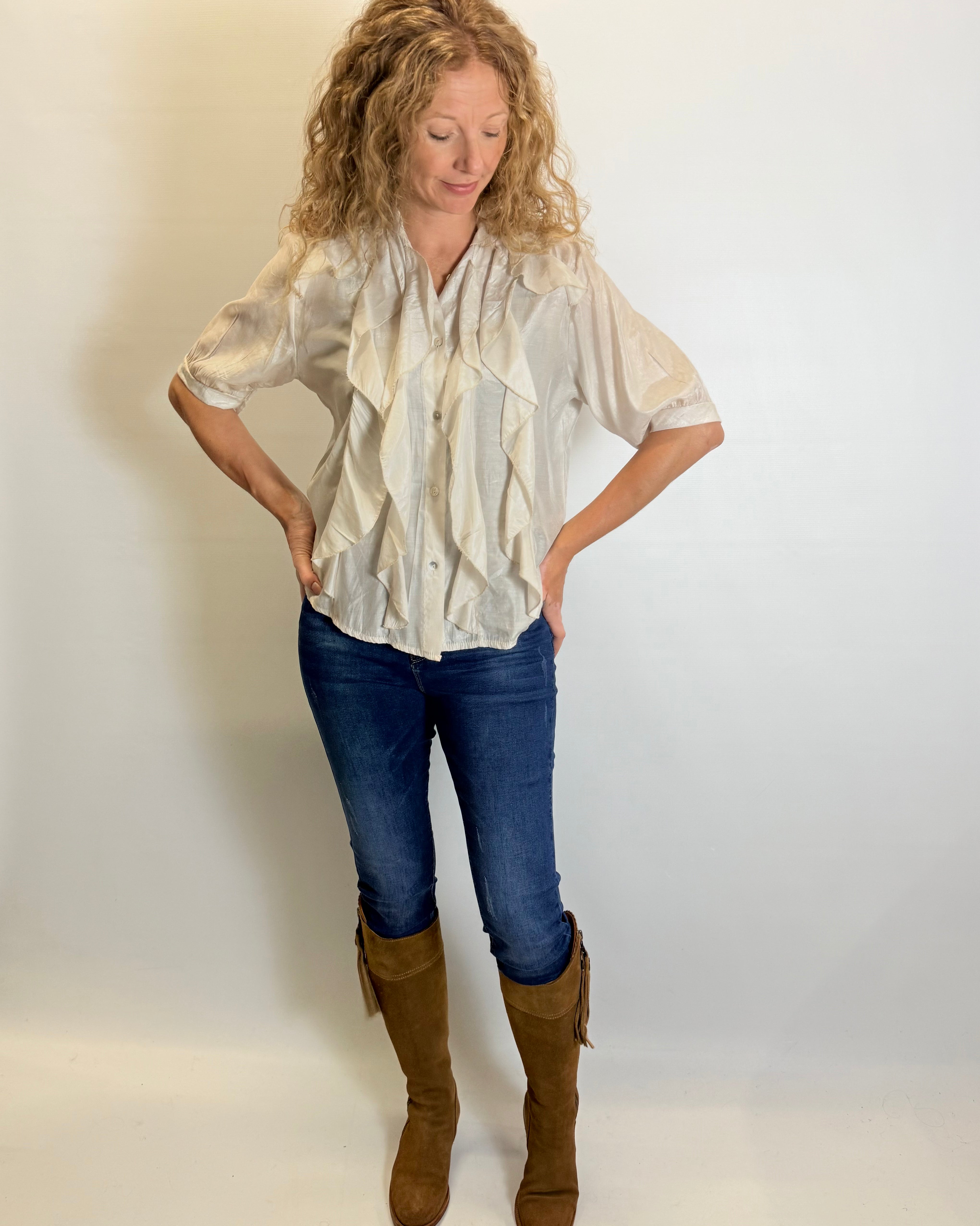 Woman wearing a white blouse, blue jeans, and brown boots against a plain background