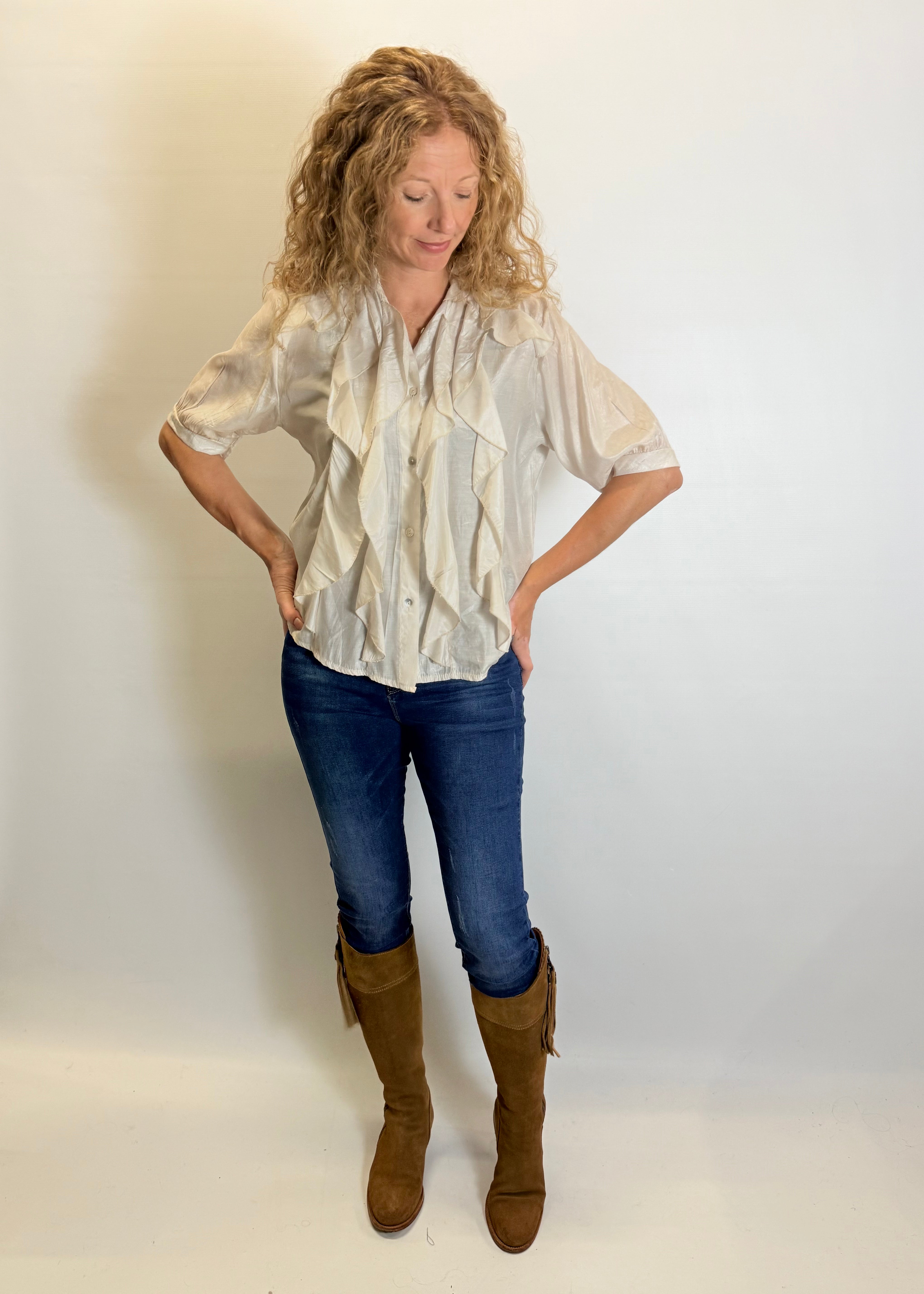 Woman wearing a white blouse, blue jeans, and brown boots against a plain background