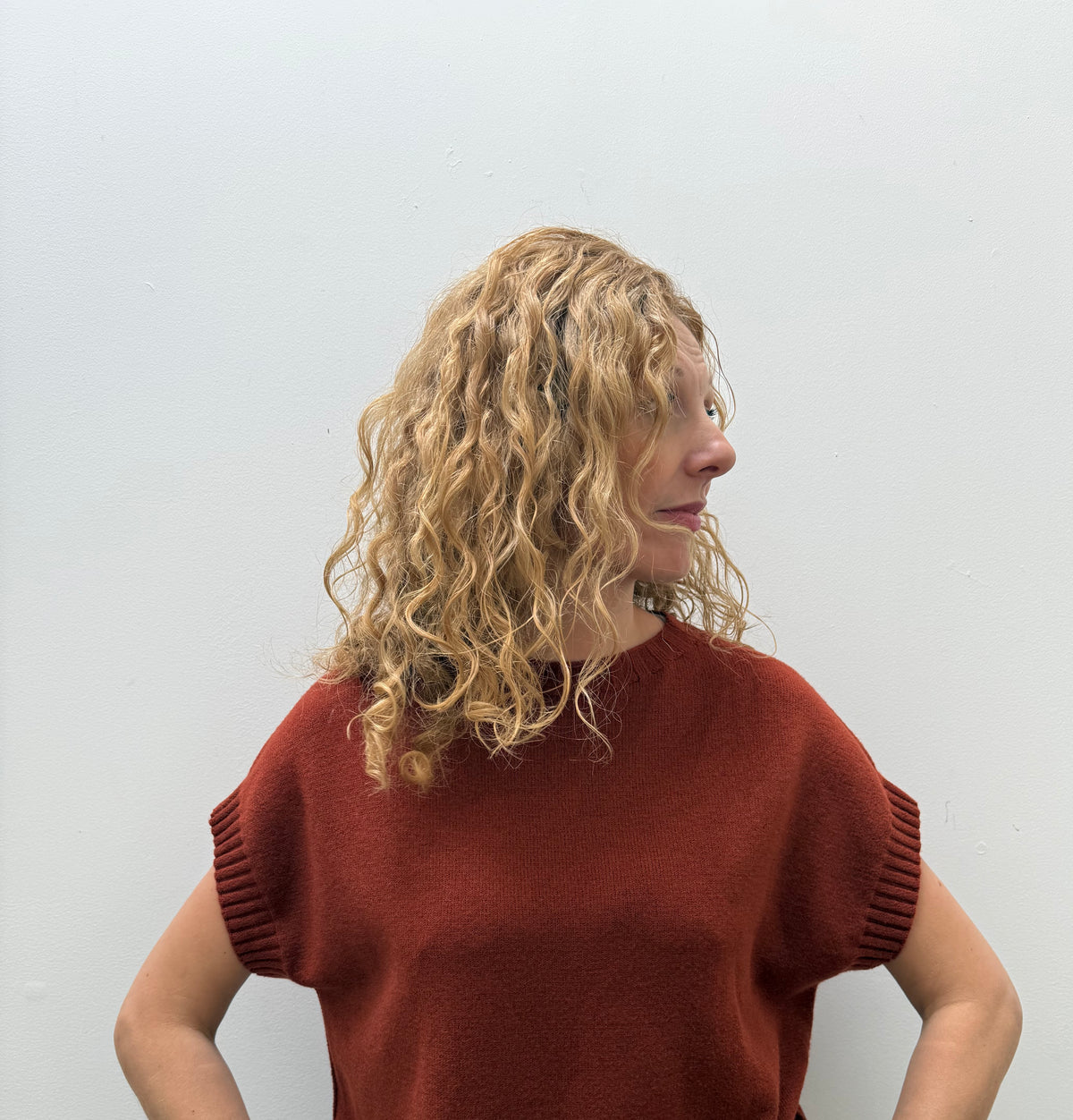 A woman with curly blonde hair wearing a rust-colored short-sleeve knit sweater and khaki pants, standing against a plain light gray wall.