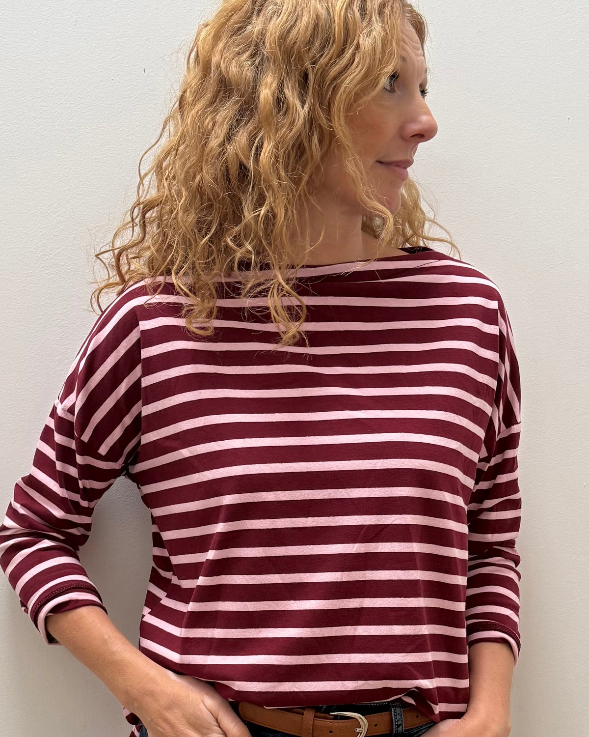 A woman with curly blonde hair wearing a burgundy and white striped long-sleeve top, dark blue jeans, and a brown belt, standing against a plain off-white wall.
