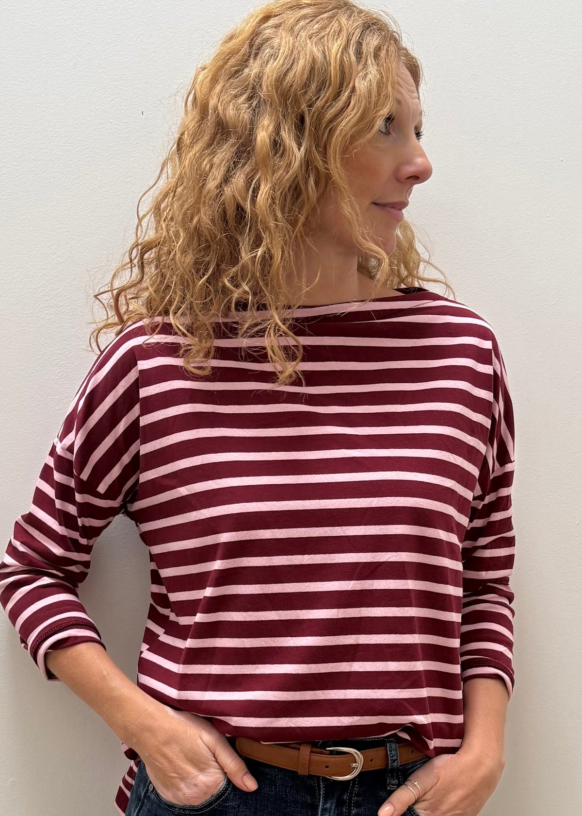 A woman with curly blonde hair wearing a burgundy and white striped long-sleeve top, dark blue jeans, and a brown belt, standing against a plain off-white wall.