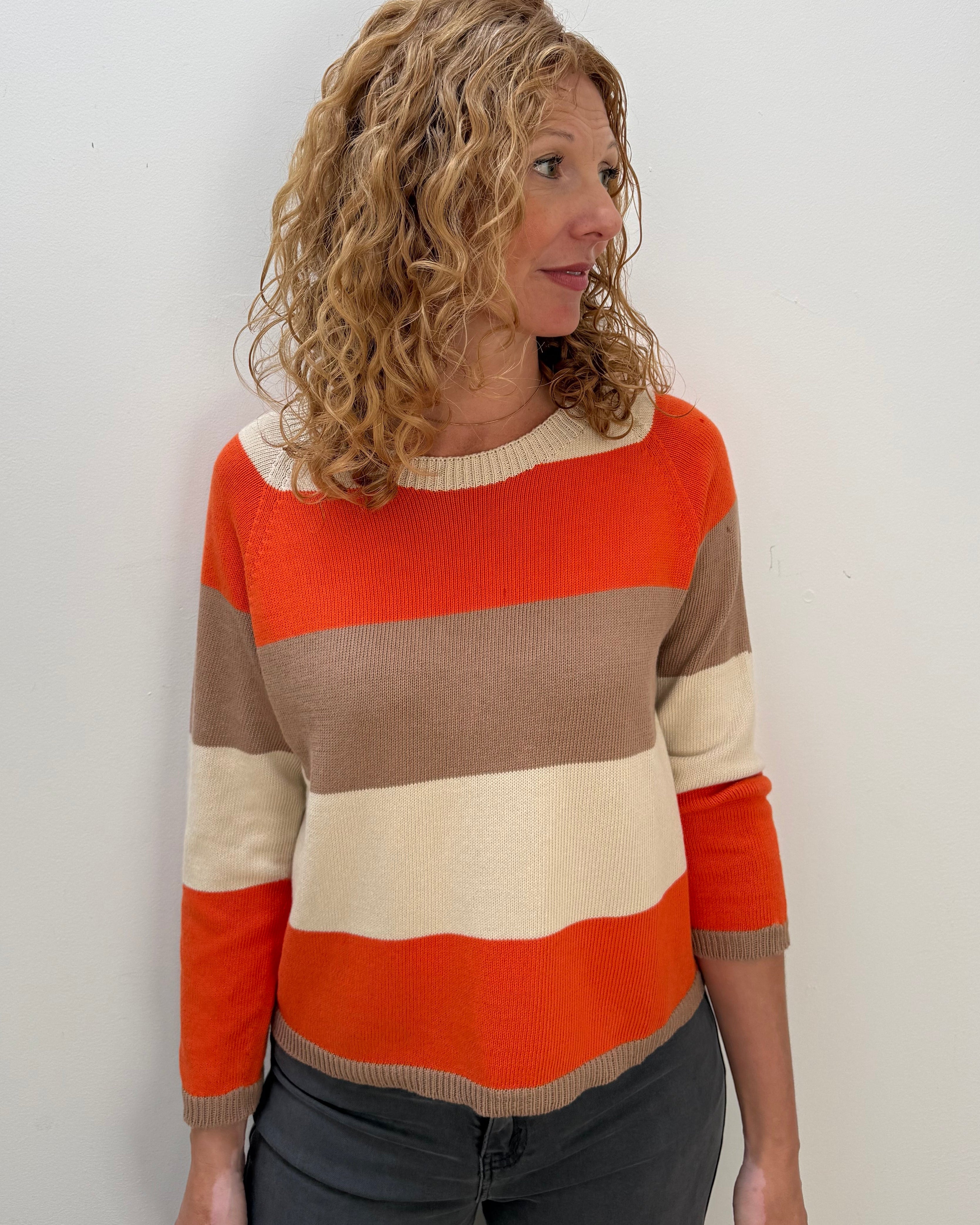A woman with curly blonde hair wearing a horizontally striped knit sweater in orange, beige, and cream, paired with dark gray pants, standing against a plain white wall.