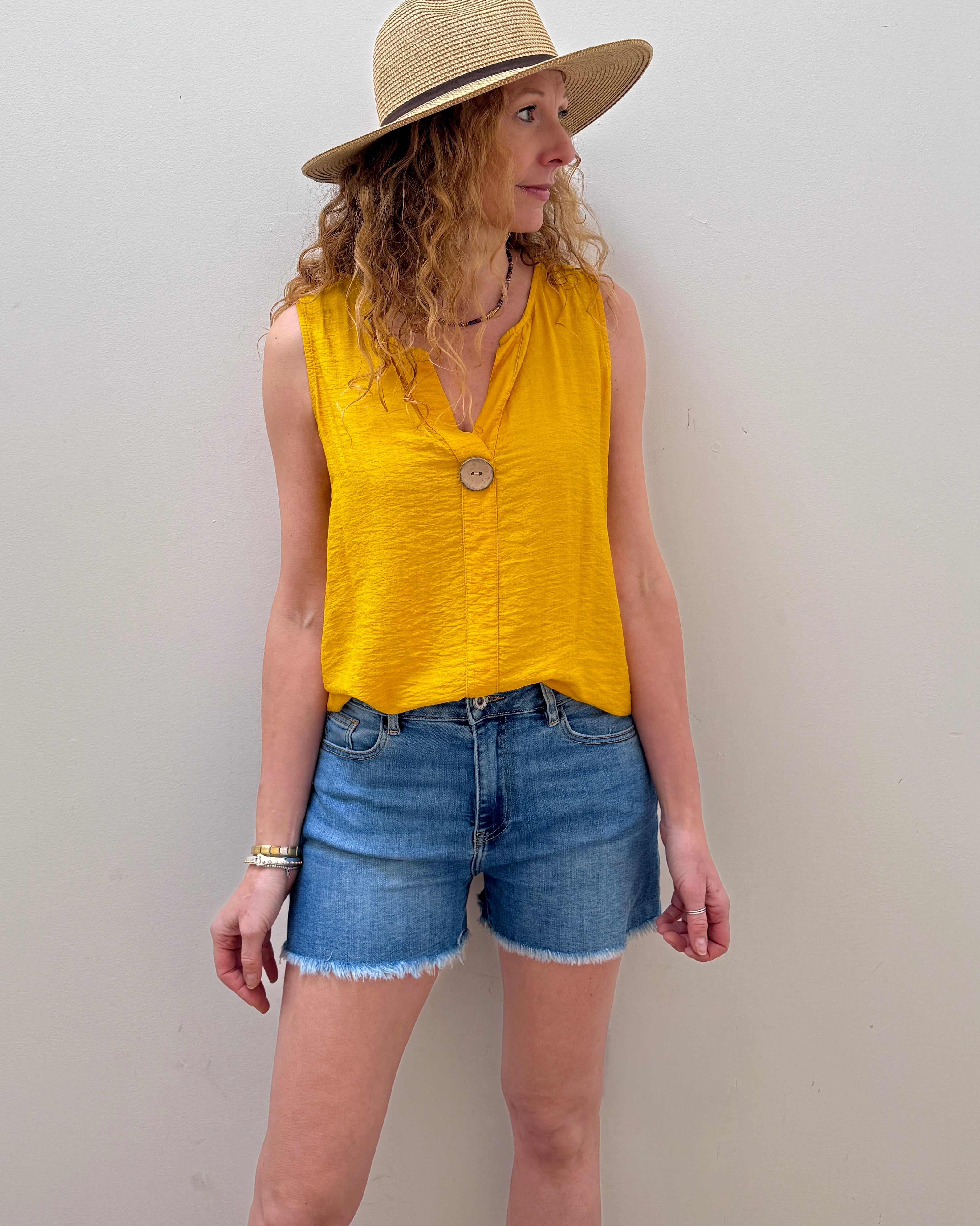 Woman wearing a yellow sleeveless top and denim shorts with a straw hat against a plain background