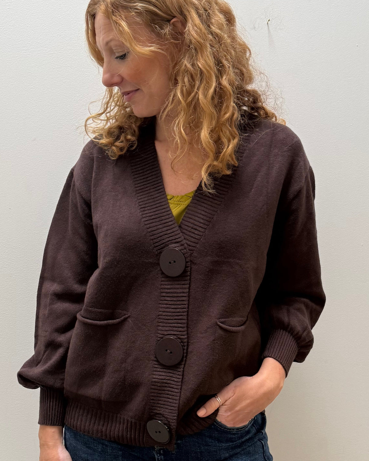 A woman wearing a dark brown button-front cardigan and blue jeans, standing in a room with a plain white wall.
