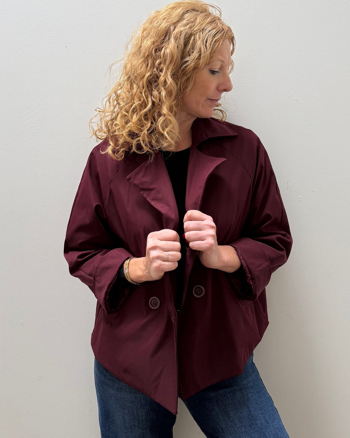 A woman with curly blonde hair wearing a dark burgundy button-up jacket and blue jeans, standing against a plain light gray wall.