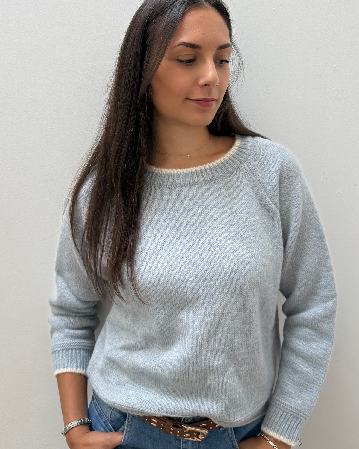 A woman wearing a light gray knit crewneck sweater, paired with blue jeans and a brown belt, standing against a plain light gray wall.