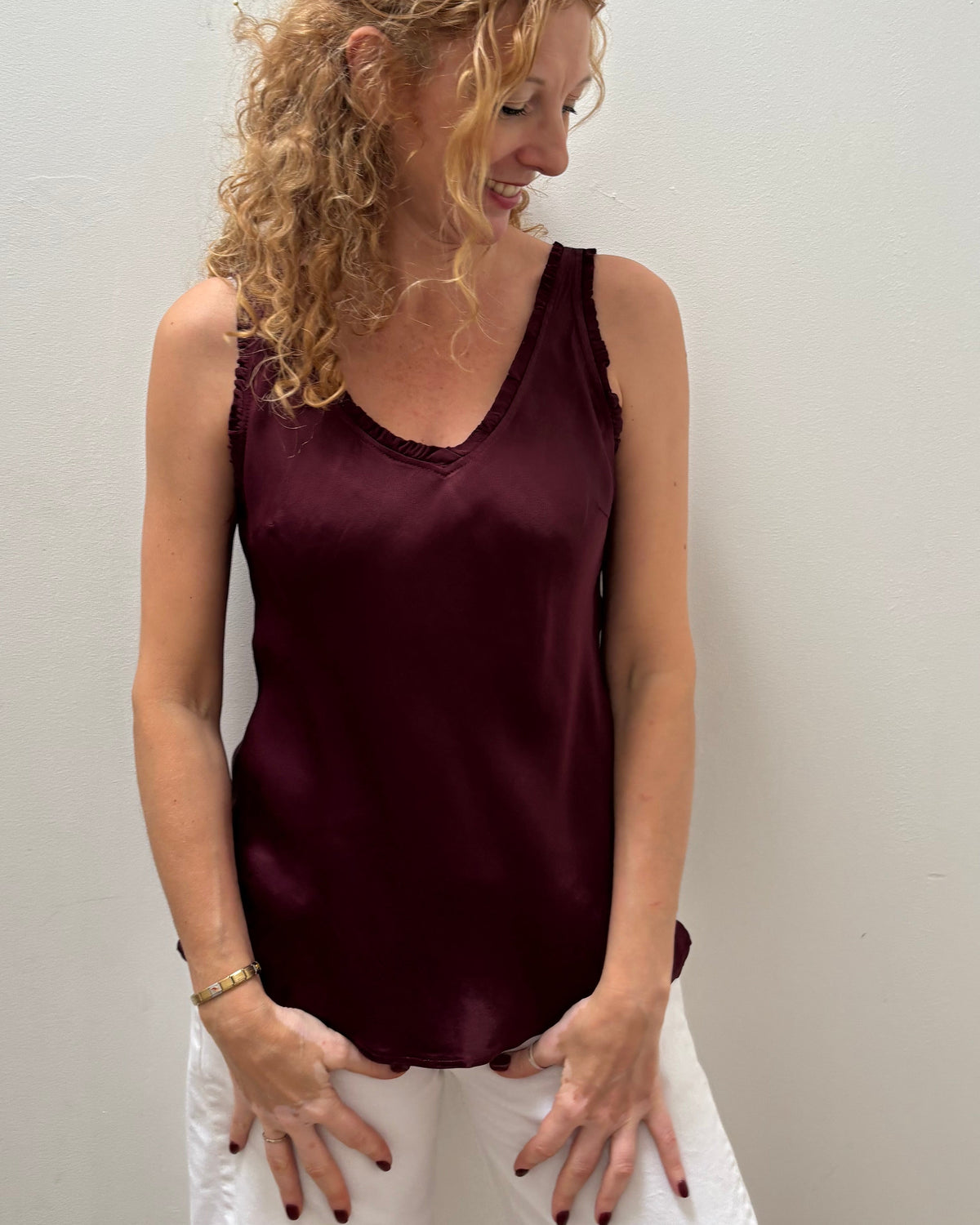 A woman with curly blonde hair wearing a sleeveless, satin-like burgundy tank top and white pants, standing against a plain off-white wall.