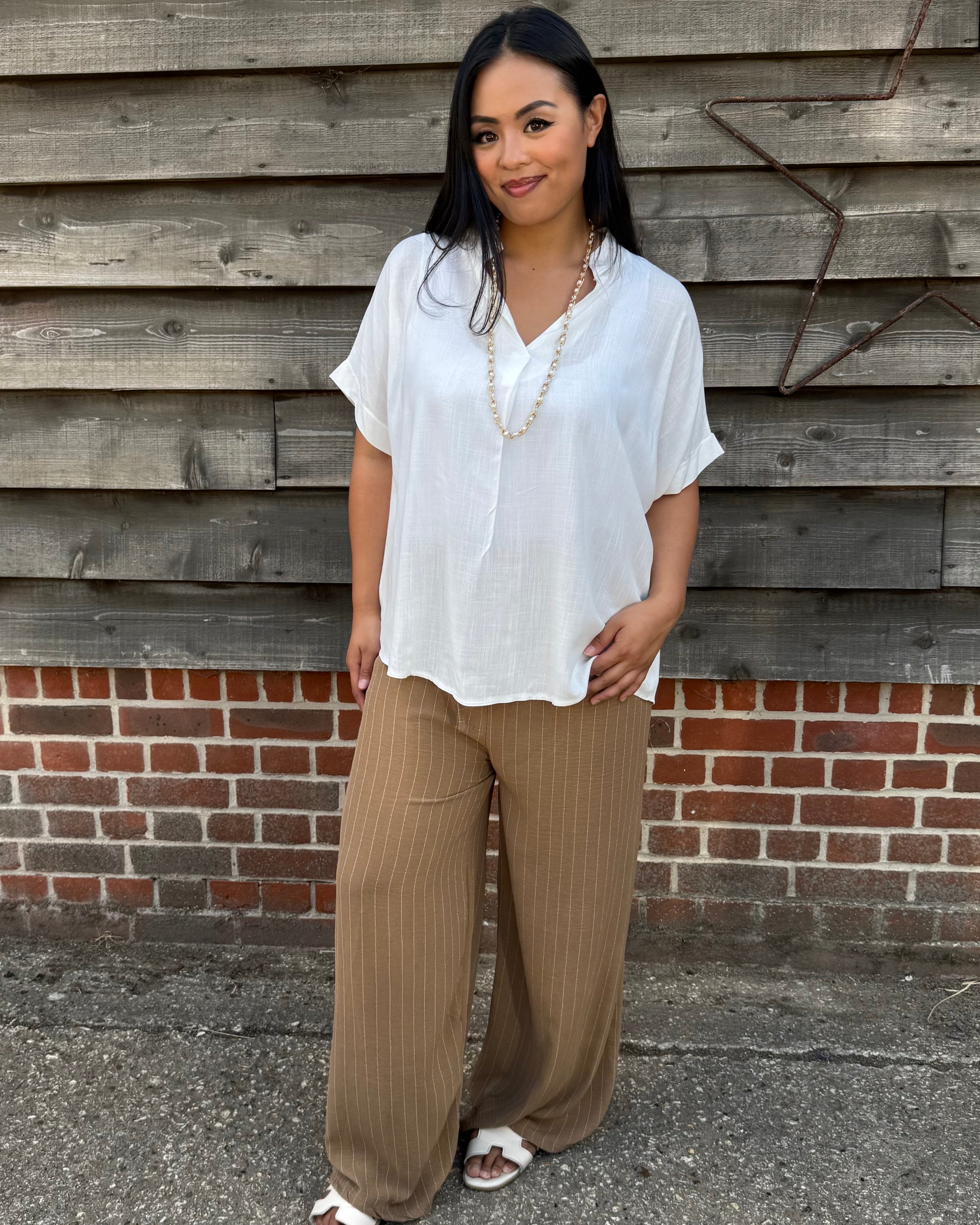 Woman wearing a white blouse and brown pants standing against a wooden wall.