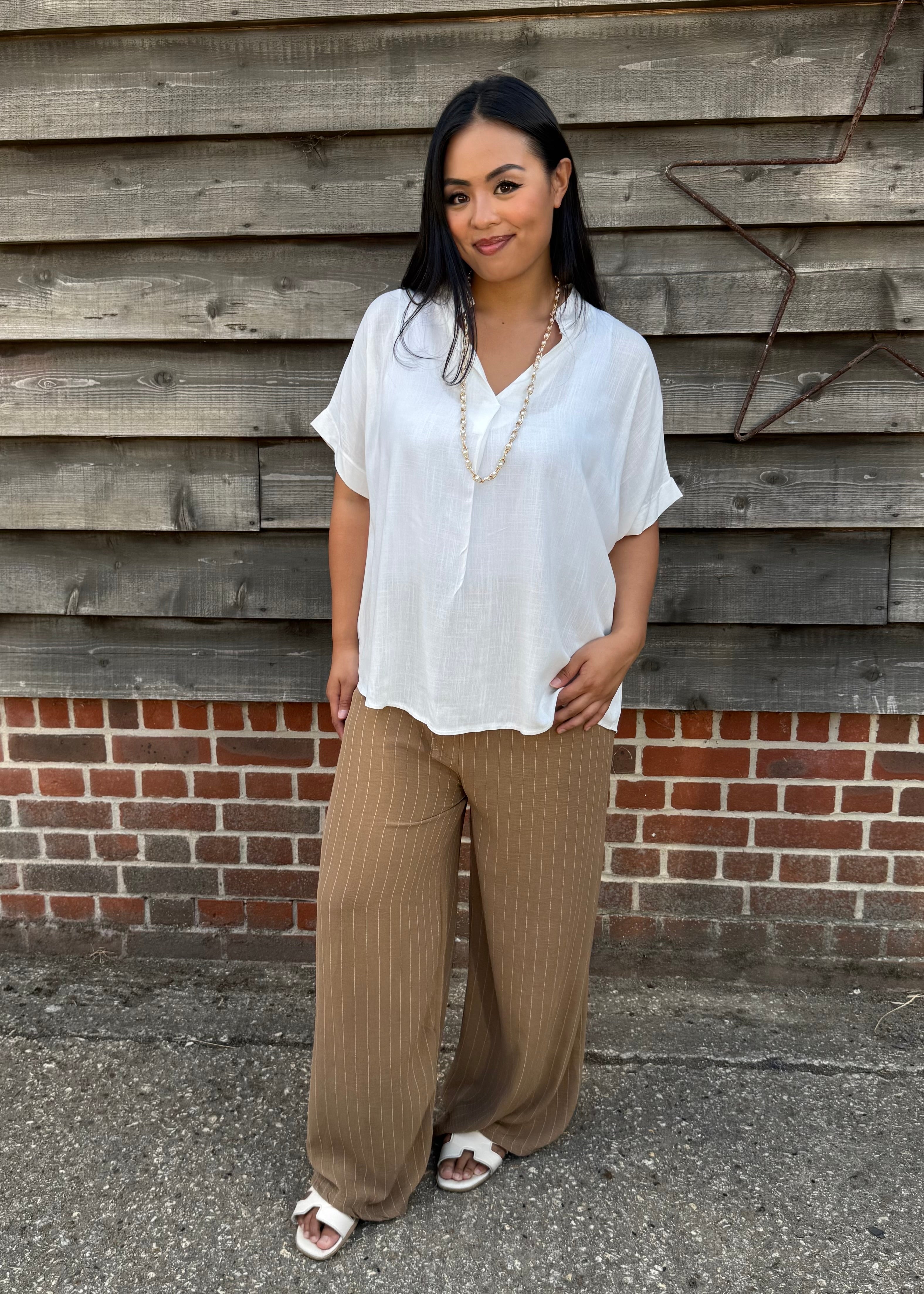 Woman wearing a white blouse and brown pants standing against a wooden wall.