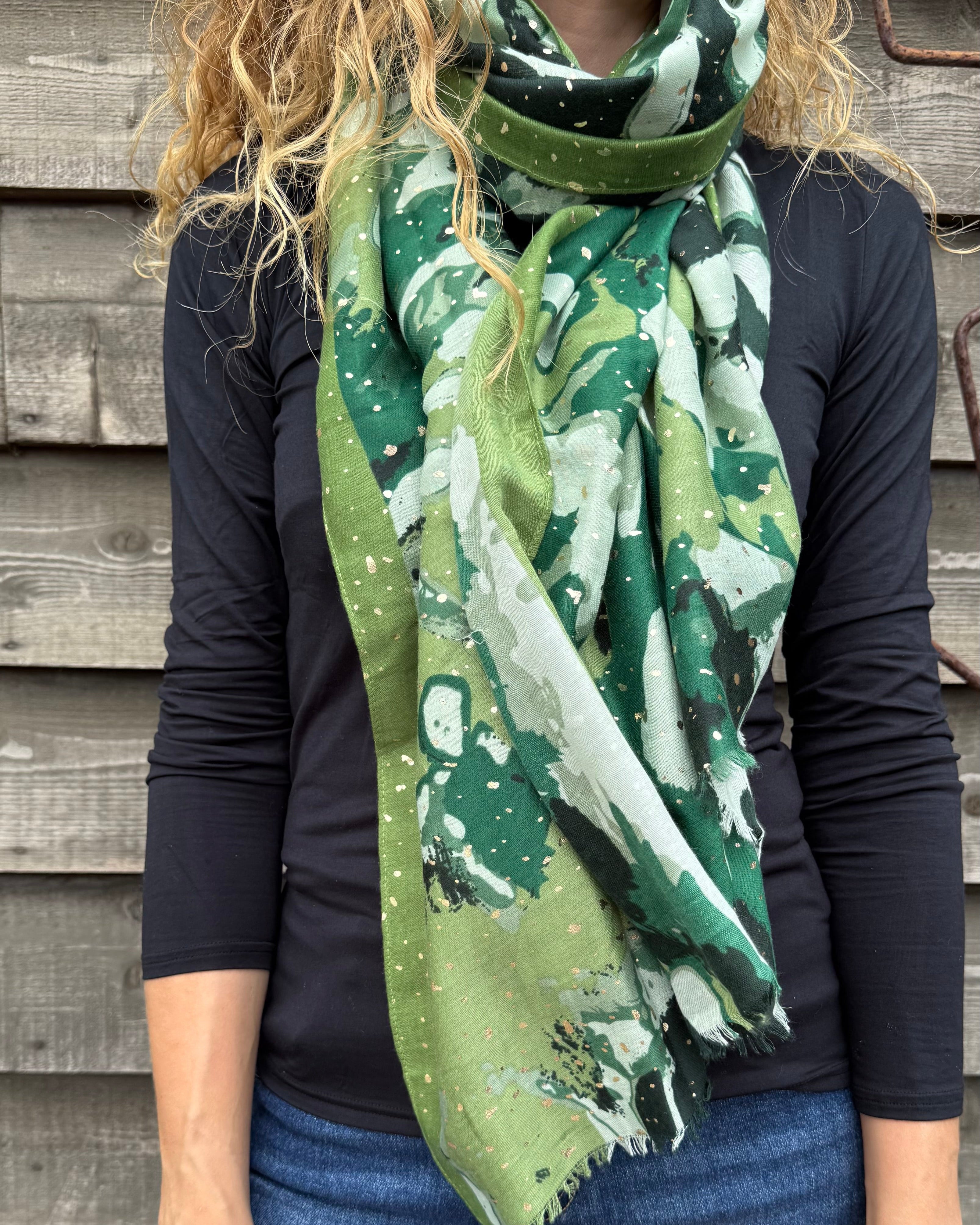 Person wearing a green patterned scarf with a dark shirt and jeans against a wooden wall.