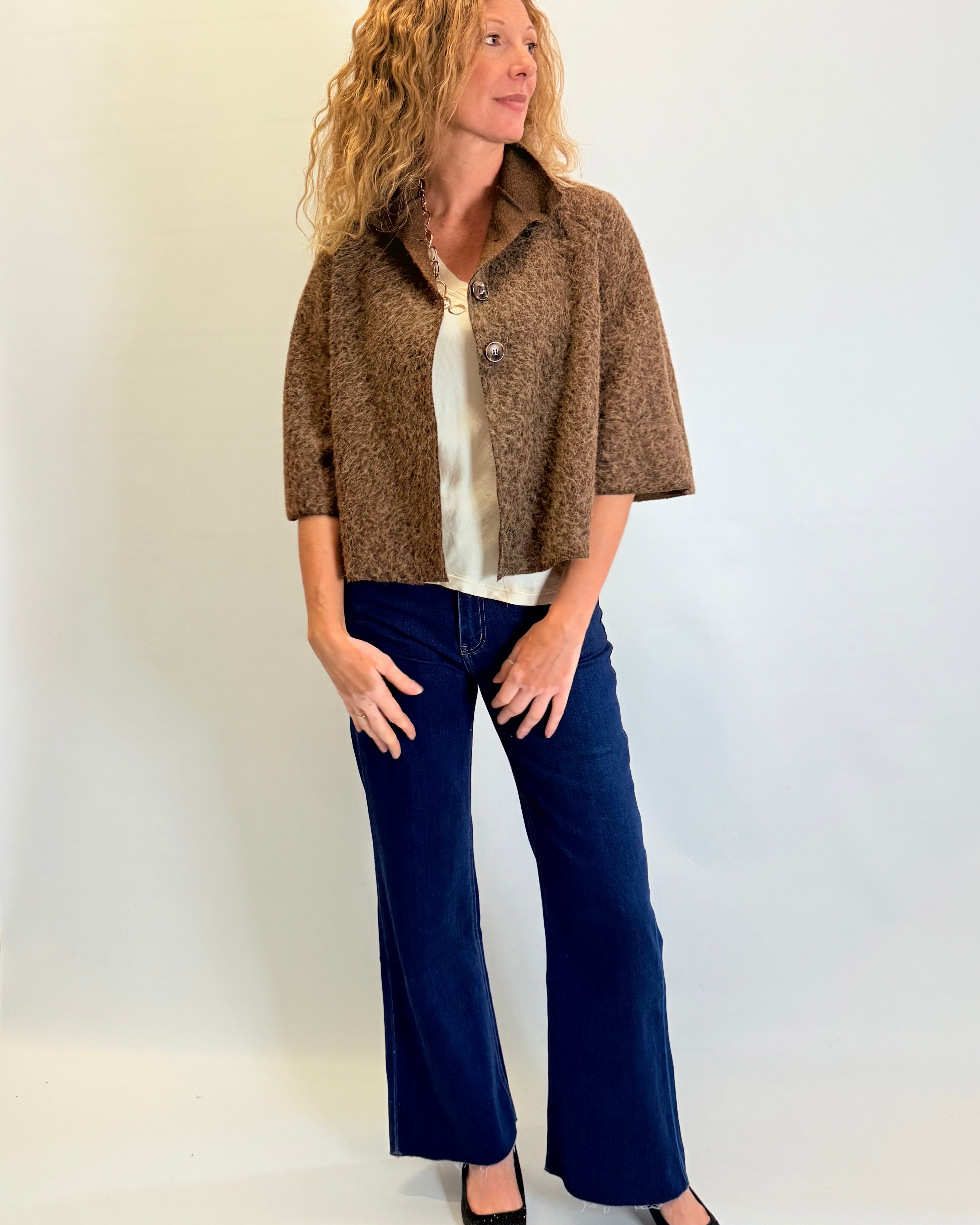 Woman wearing a brown patterned jacket, white blouse, and blue jeans against a plain background