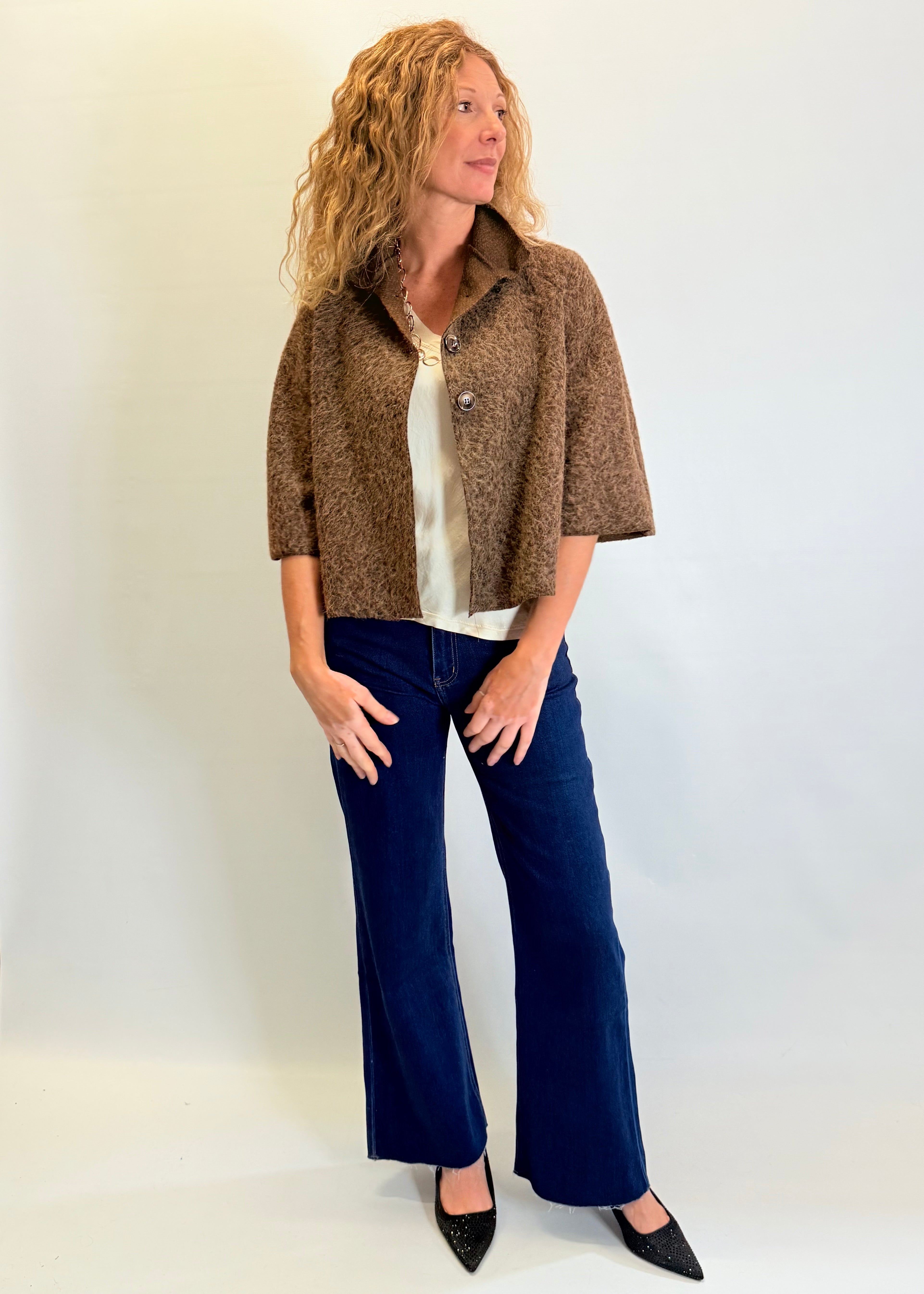 Woman wearing a brown patterned jacket, white blouse, and blue jeans against a plain background