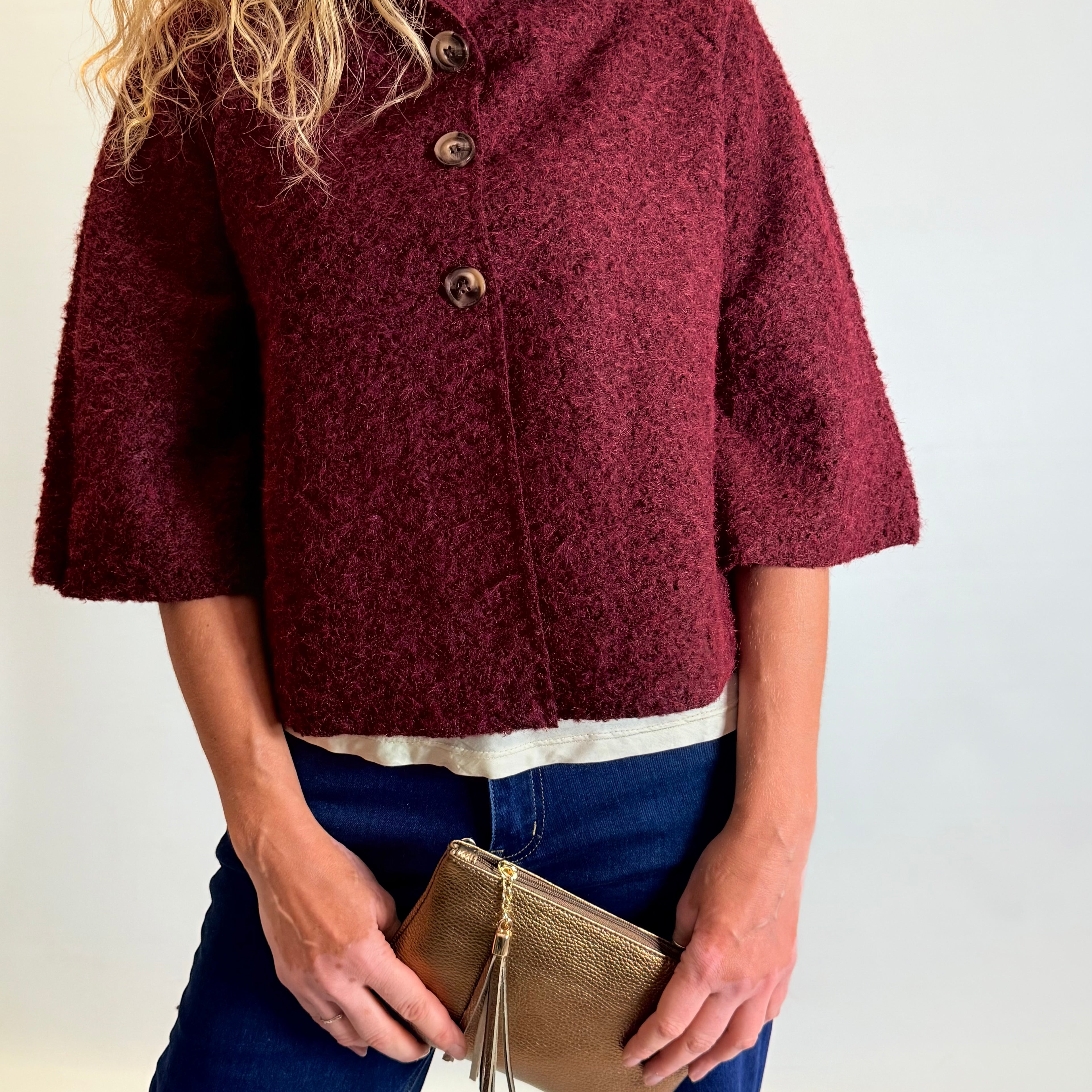 Person wearing a burgundy textured jacket holding a gold clutch against a white background