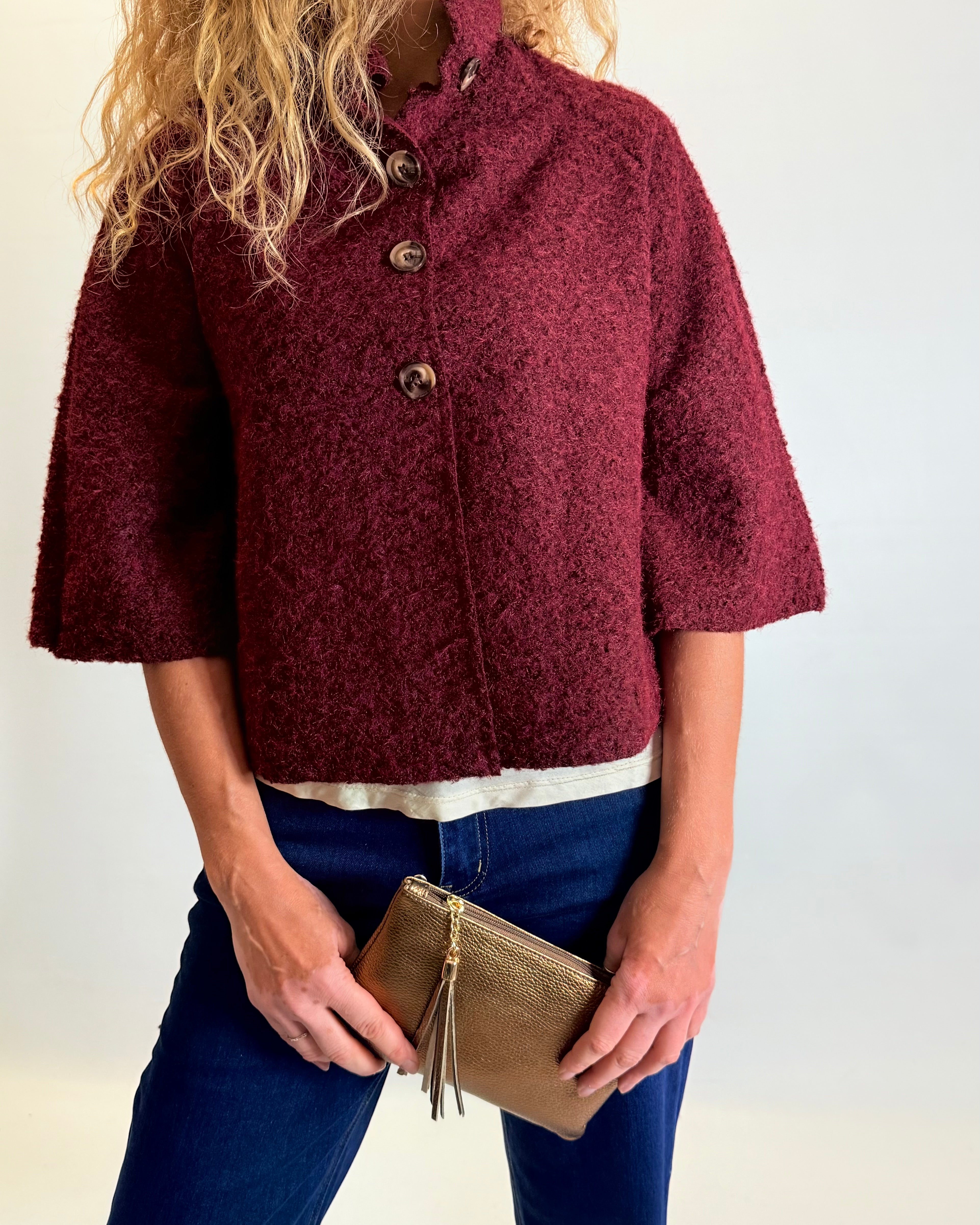 Person wearing a burgundy textured jacket holding a gold clutch against a white background