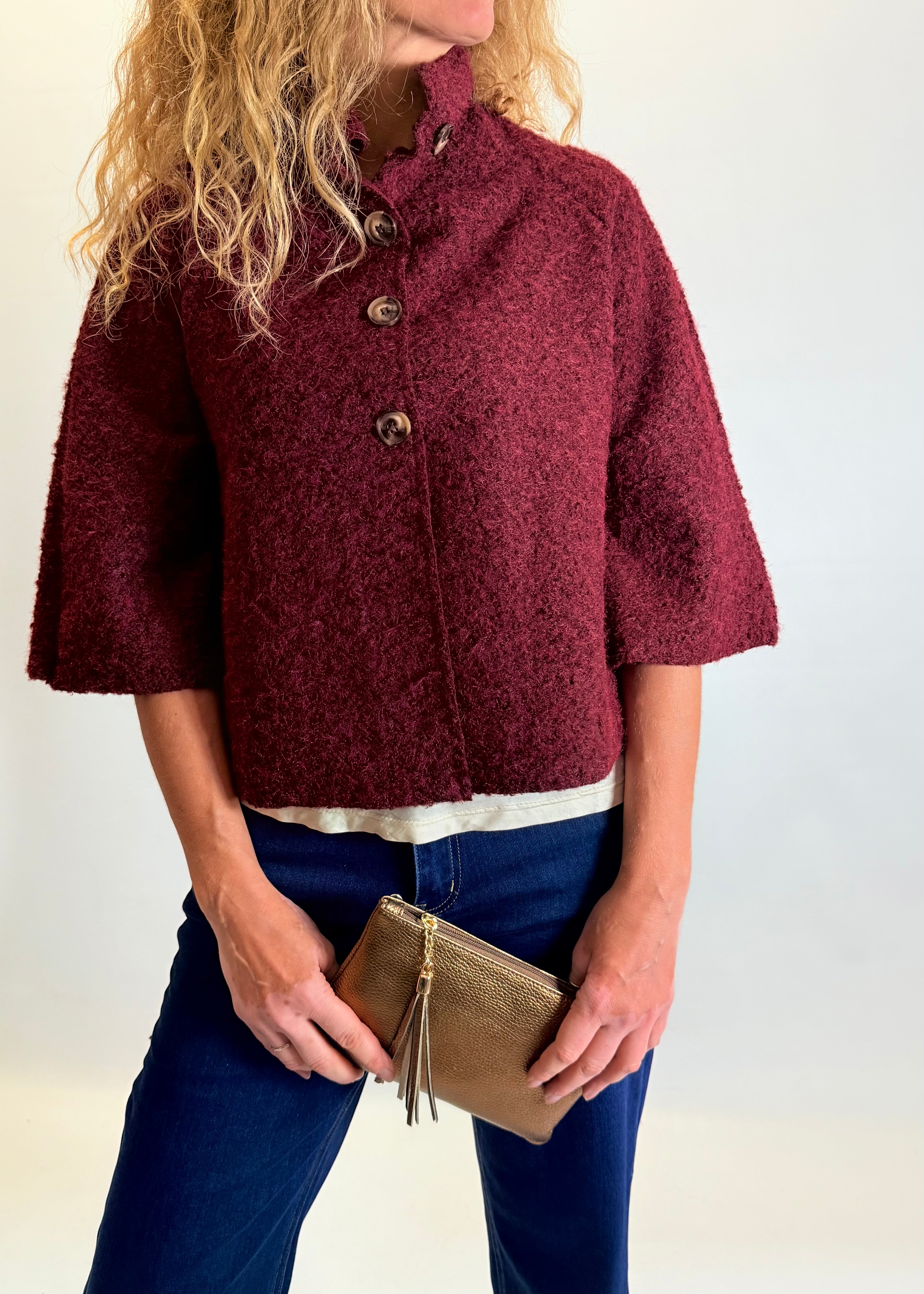 Person wearing a burgundy textured jacket holding a gold clutch against a white background